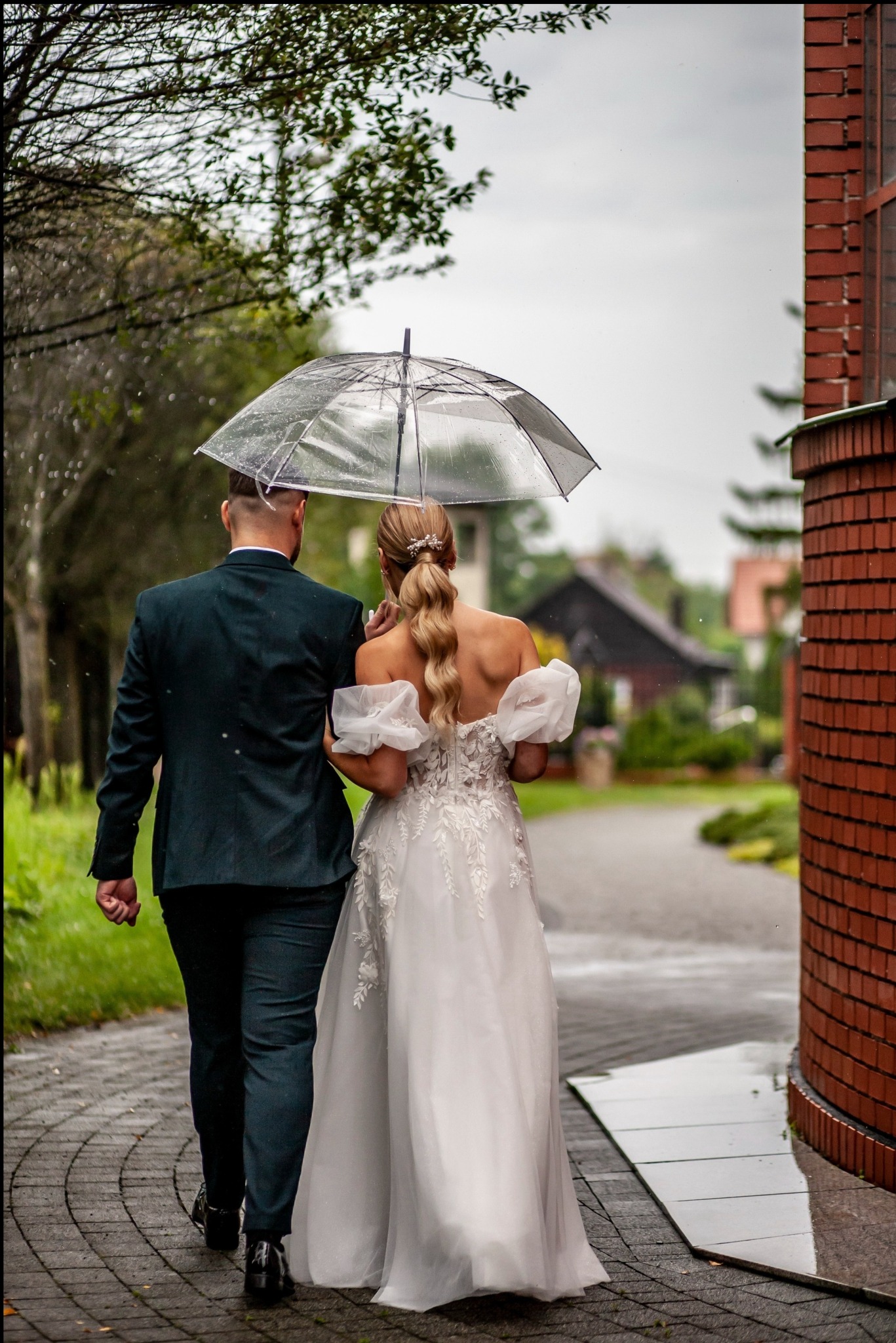 Młoda para w deszczu idzie ścieżką pod przezroczystym parasolem. Panna młoda w białej sukni z odkrytymi plecami, pan młody w ciemnozielonym garniturze.