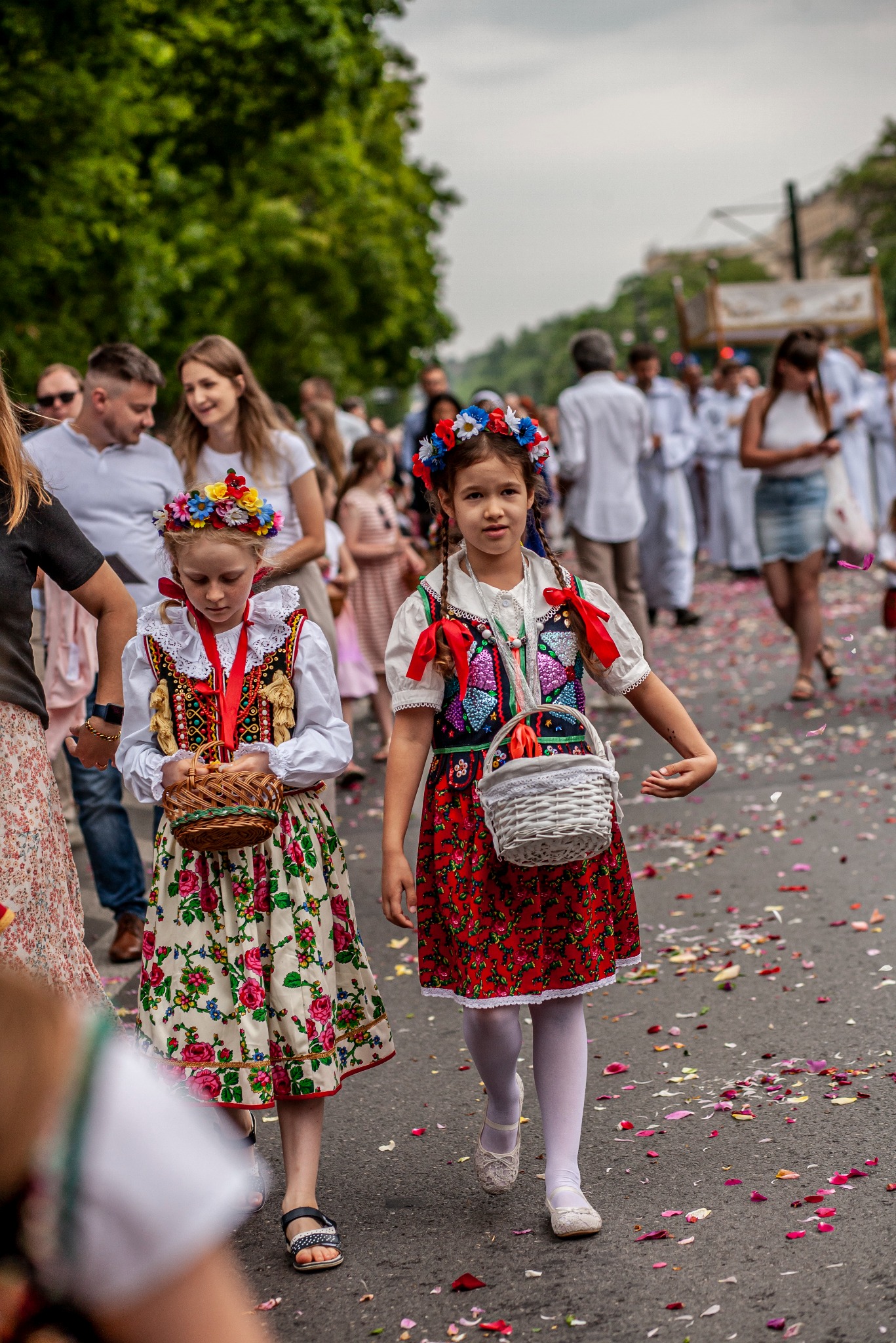 Dwie dziewczynki w barwnych, ludowych strojach z wiankami na głowach, niosące koszyczki, uczestniczą w procesji. Tło stanowią inni uczestnicy i miejska sceneria.
