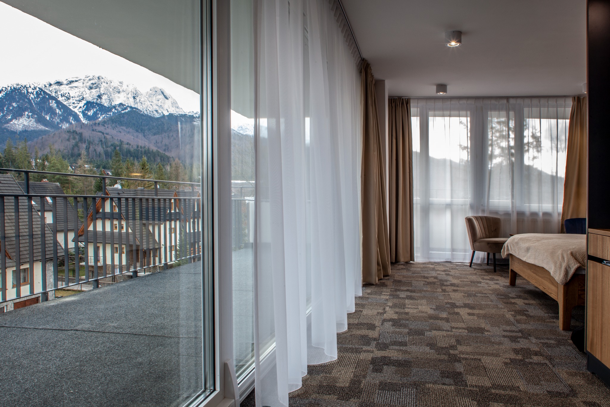 Jasny pokój hotelowy z balkonem i widokiem na góry. Przez okno widać ośnieżone szczyty i zabudowę. Wnętrze z beżowymi zasłonami i dywanem w geometryczne wzory.