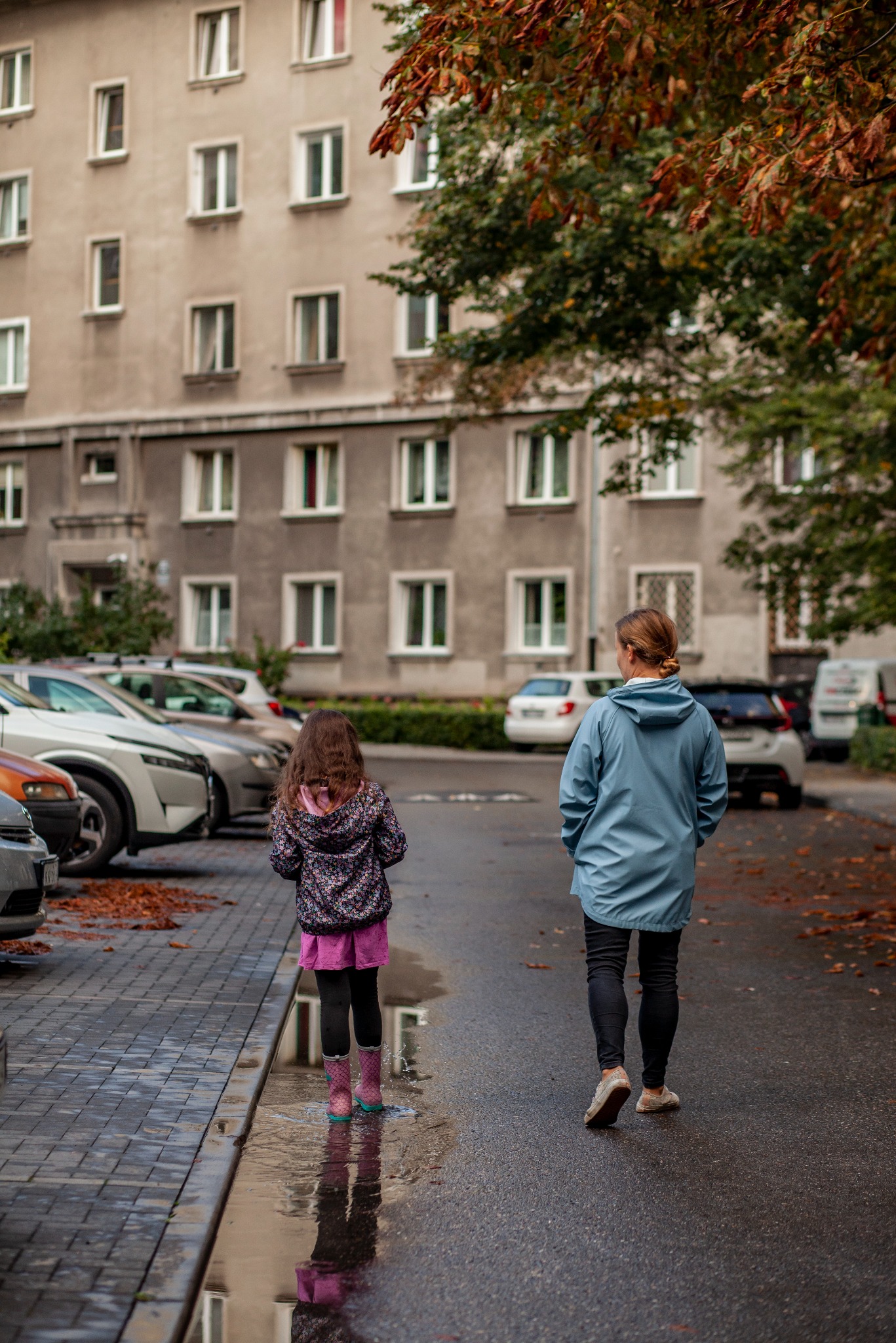 Jesienne popołudnie: Dziewczynka w kaloszach i kobieta spacerują po mokrej ulicy, odbicie w kałuży, blok mieszkalny w tle, opadłe liście.