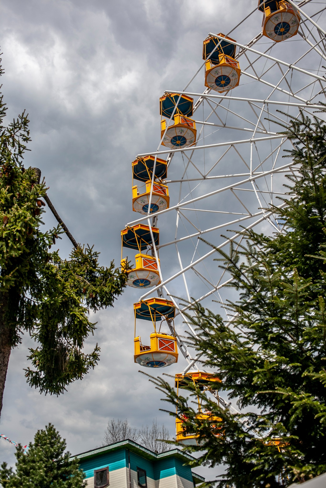 Żółto-białe koło młyńskie z kabinami, widziane z dołu, częściowo zasłonięte przez drzewa iglaste na tle pochmurnego nieba. Widoczny fragment budynku.