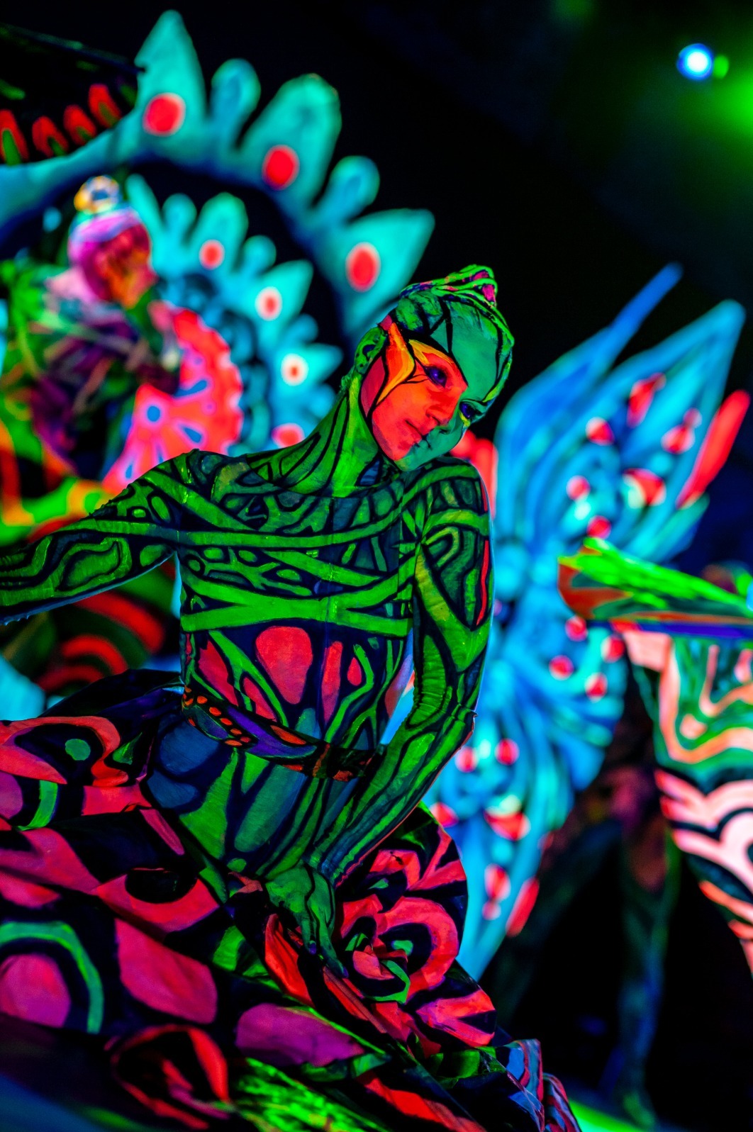 Artystyczne zdjęcie kobiety z fluorescencyjnym body paintingiem w odcieniach zieleni, różu i błękitu, na tle dekoracji scenicznej w podobnej stylistyce, oświetlonej światłem UV.