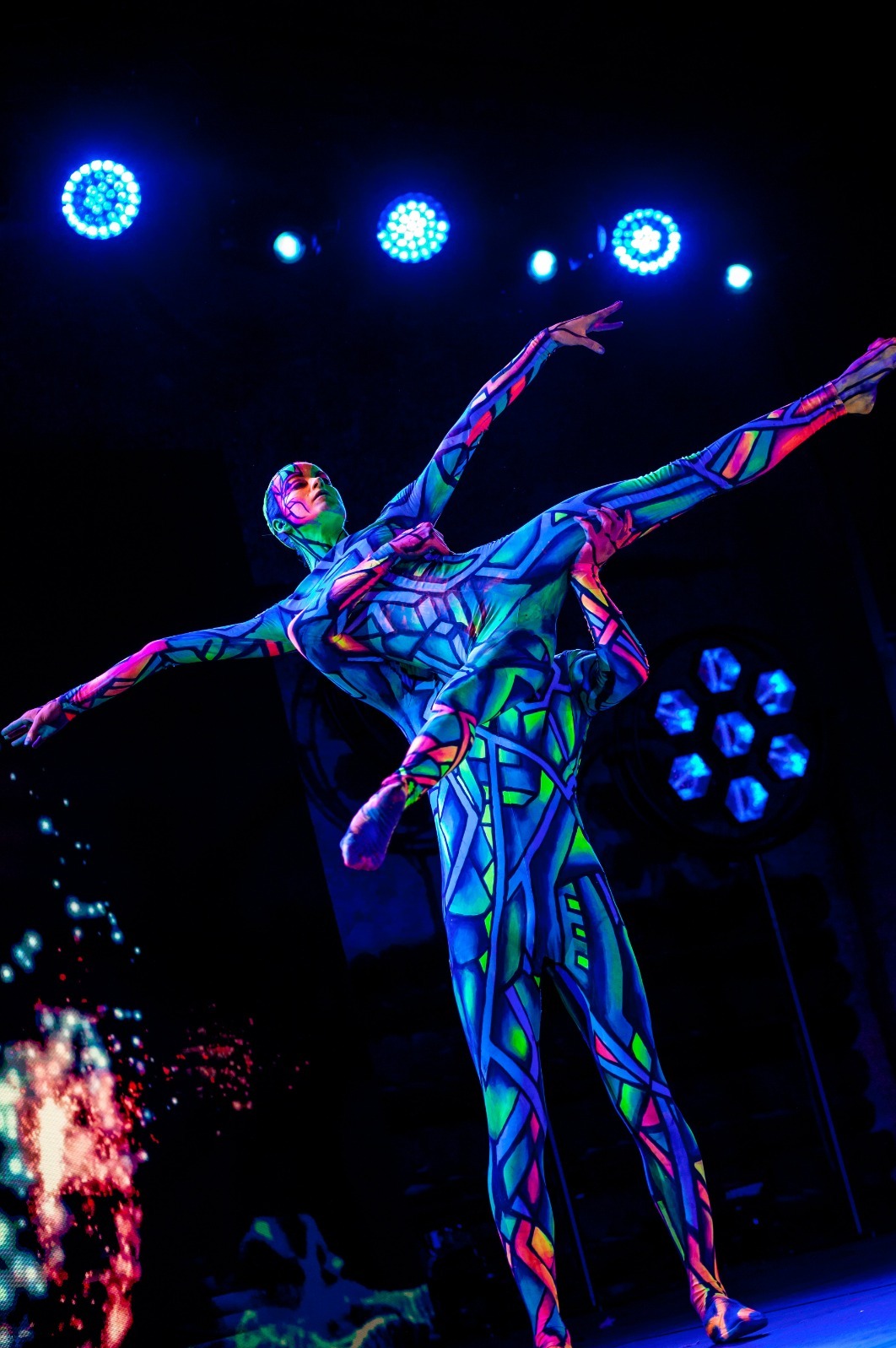 Akrobatyczny duet w neonowych kostiumach, podświetlony niebieskimi reflektorami, prezentuje dynamiczną figurę podczas występu na scenie.