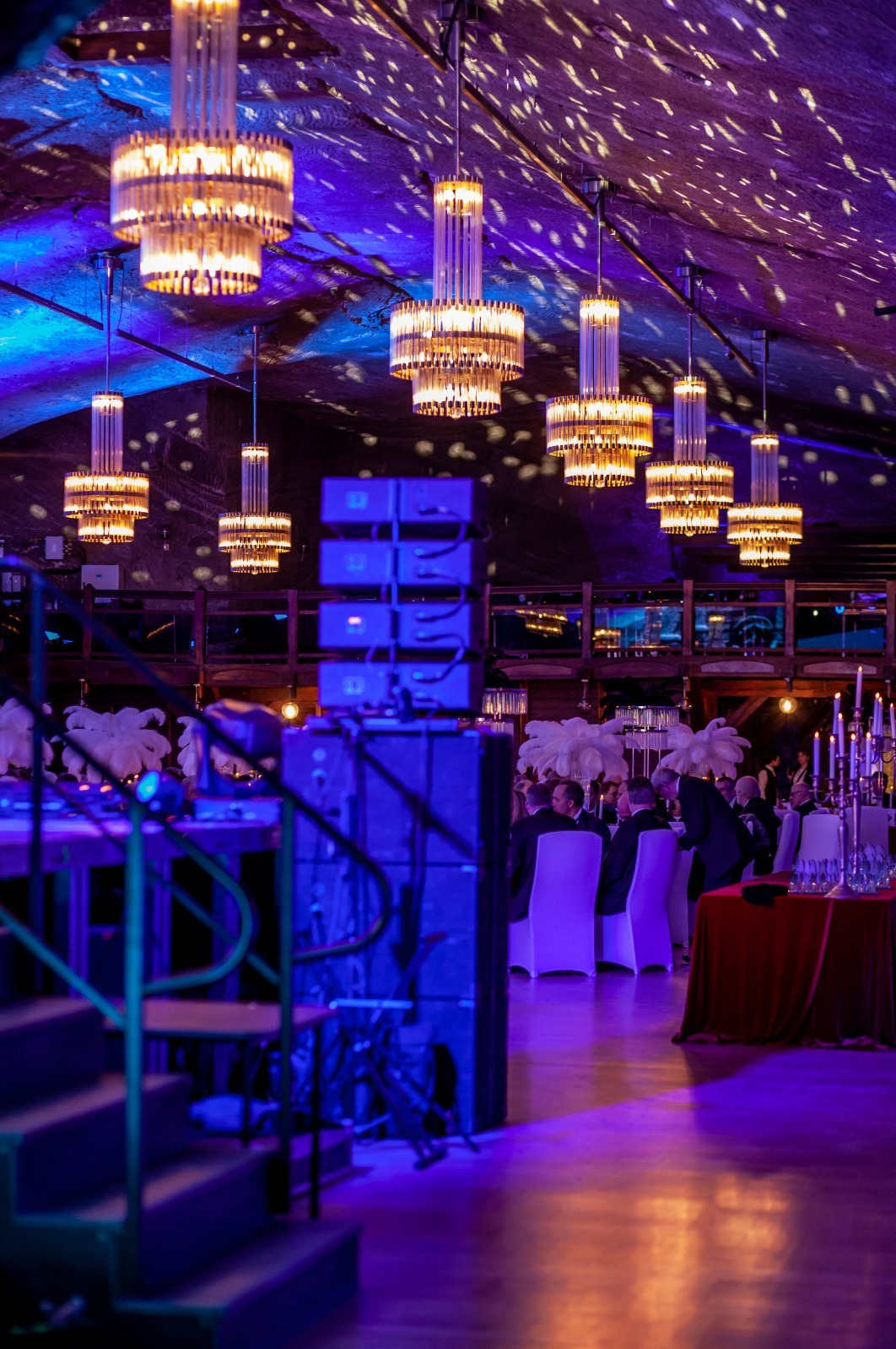 Eleganckie przyjęcie w sali z kryształowymi żyrandolami i nastrojowym, niebiesko-fioletowym oświetleniem. Widoczne stoliki z gośćmi i sprzęt nagłośnieniowy.
