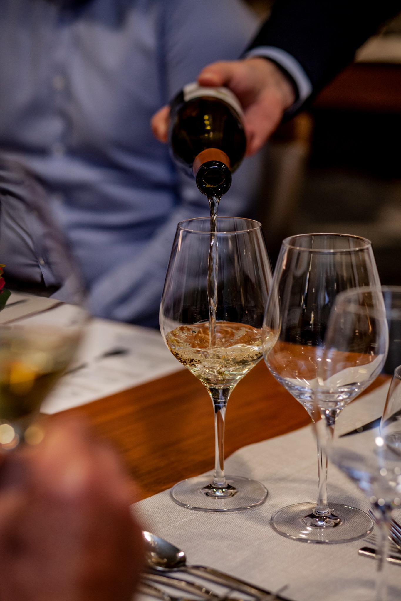 Nalewanie białego wina z butelki do kieliszka na stole z zastawą. Ujęcie z bliska, skupione na płynie i szkle, z rozmytym tłem i elementami stroju osoby nalewającej.