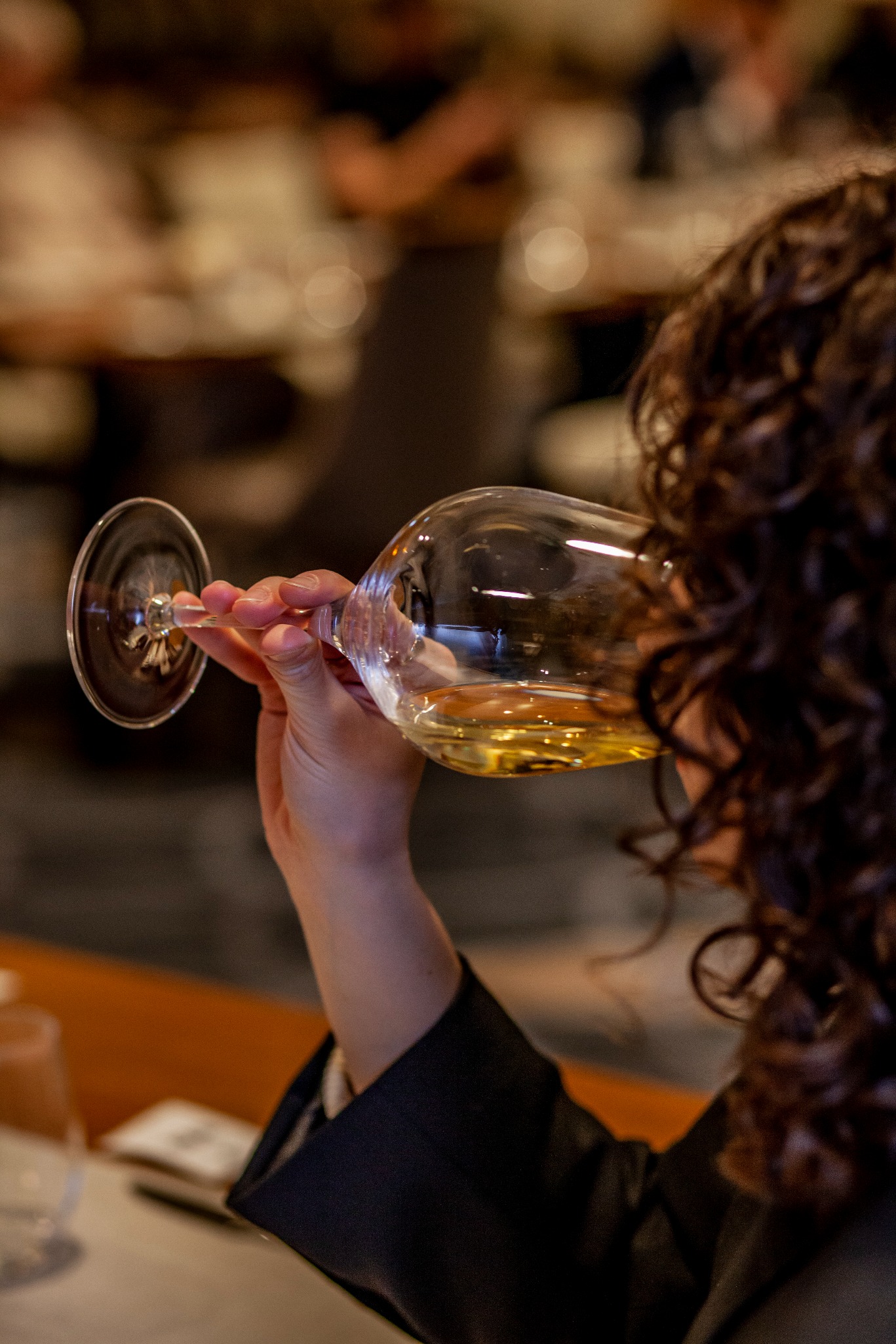 Osoba z kręconymi włosami pije białe wino z kieliszka na tle rozmytego wnętrza restauracji. Widoczna dłoń trzymająca kieliszek i fragment czarnej marynarki.