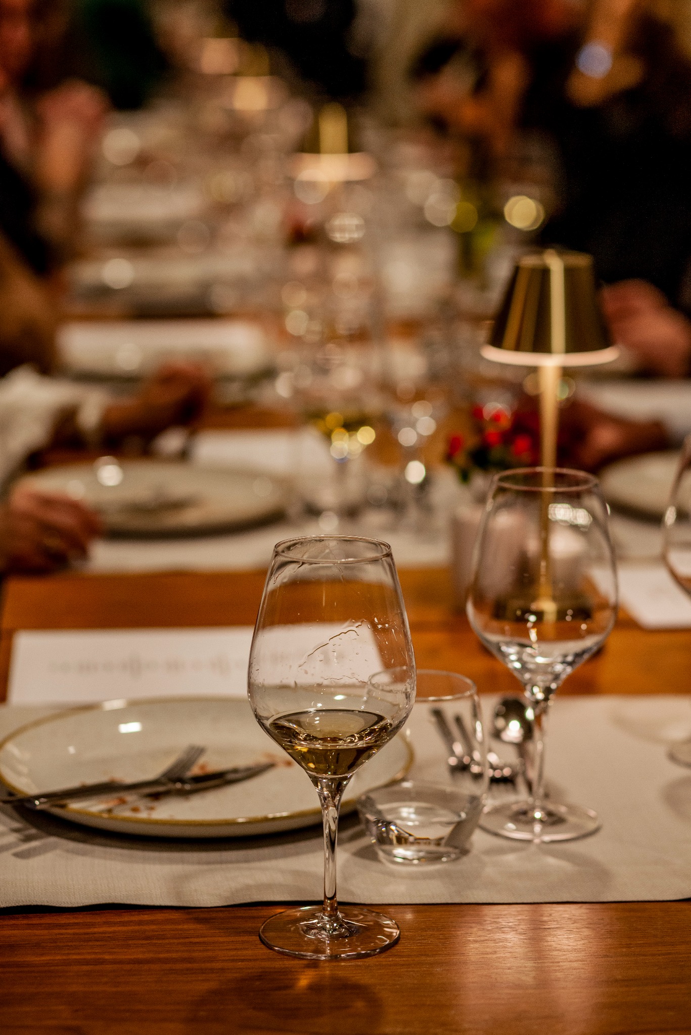 Ustawiony stół z kieliszkami wina, talerzami i sztućcami, oświetlony ciepłym światłem lampki, sugerujący eleganckie przyjęcie lub uroczystą kolację.