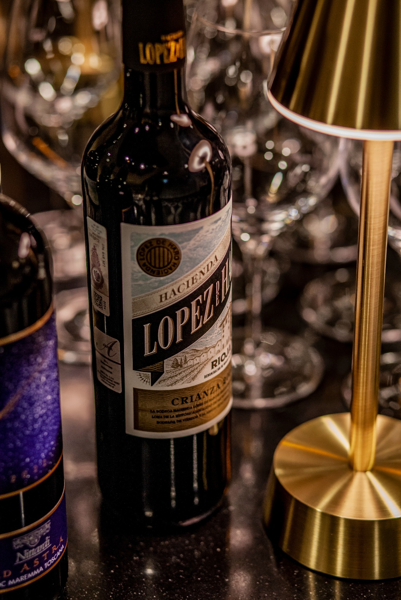 Butelka wina Rioja Crianca Hacienda Lopez de Haro, oświetlona złotą lampką, na tle kieliszków. Etykieta z detalami, w tle rozmyte szkło.