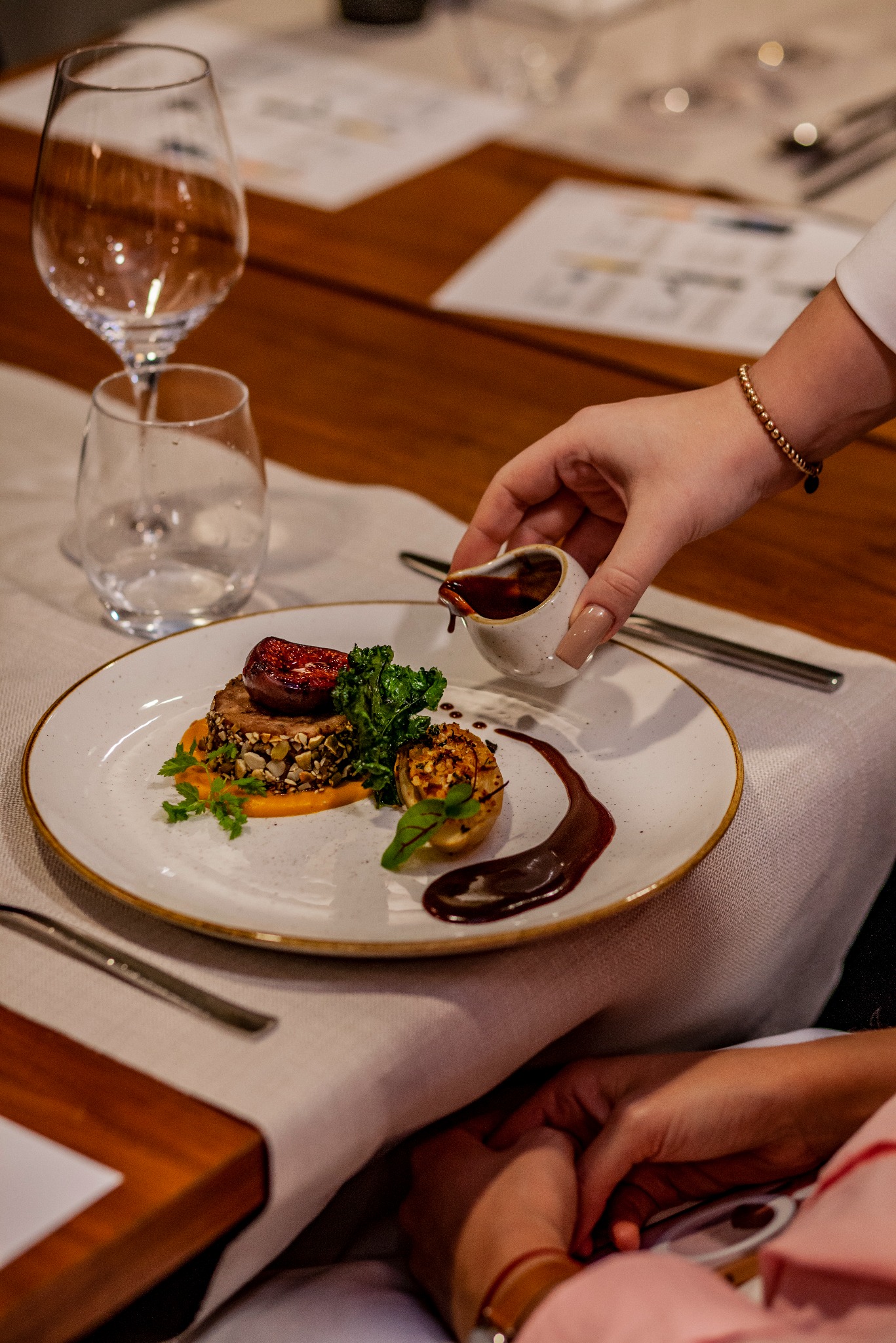 Elegancko podane danie z figą i sosem na białym talerzu, z ręką polewającą sosem z dzbanka. W tle kieliszki i drewniany stół. Fine dining.