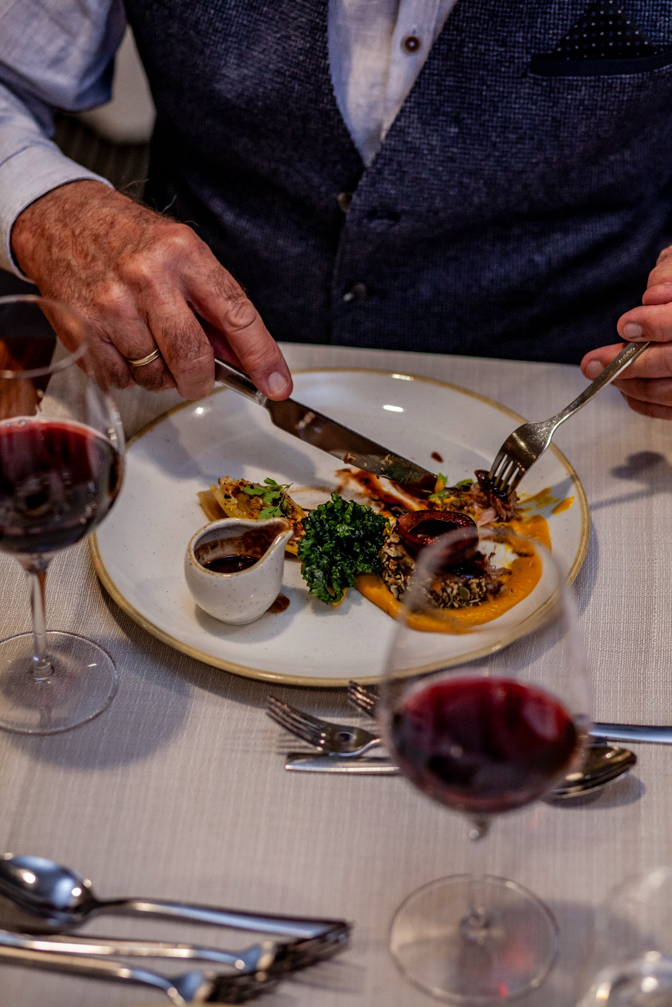 Elegancko podane danie na talerzu, krojone nożem i widelcem przez mężczyznę w kamizelce, z kieliszkami czerwonego wina w tle. Fine dining w restauracji.