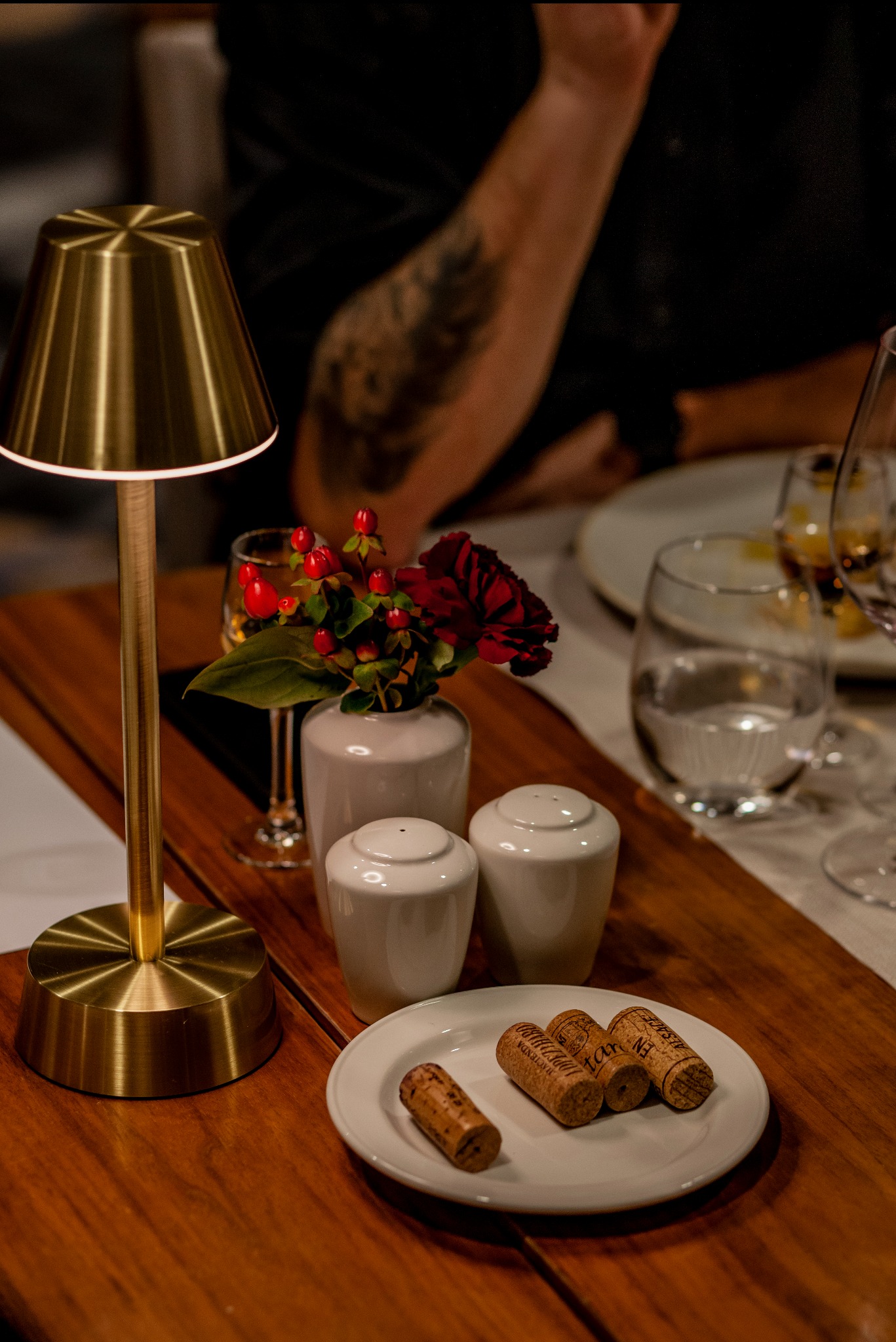 Ustawienie stołu z lampką, korkami po winie na talerzu, kwiatami w wazonie i przyprawnikami. Ciepłe światło, elegancka aranżacja.