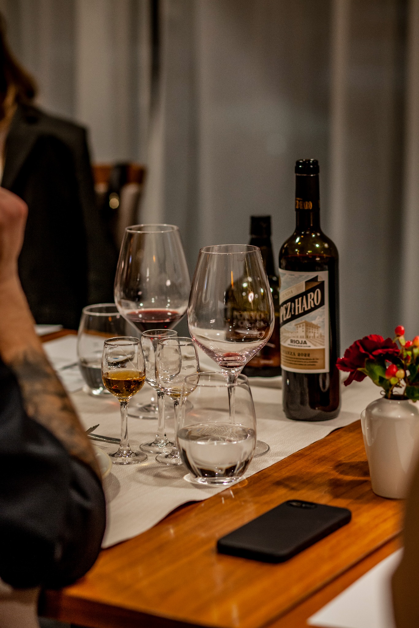 Ustawiony stół z butelką wina Pez de Haro Rioja, kieliszkami z winem i wodą, oraz smartfonem na drewnianym blacie. W tle zasłony i fragmenty osób.