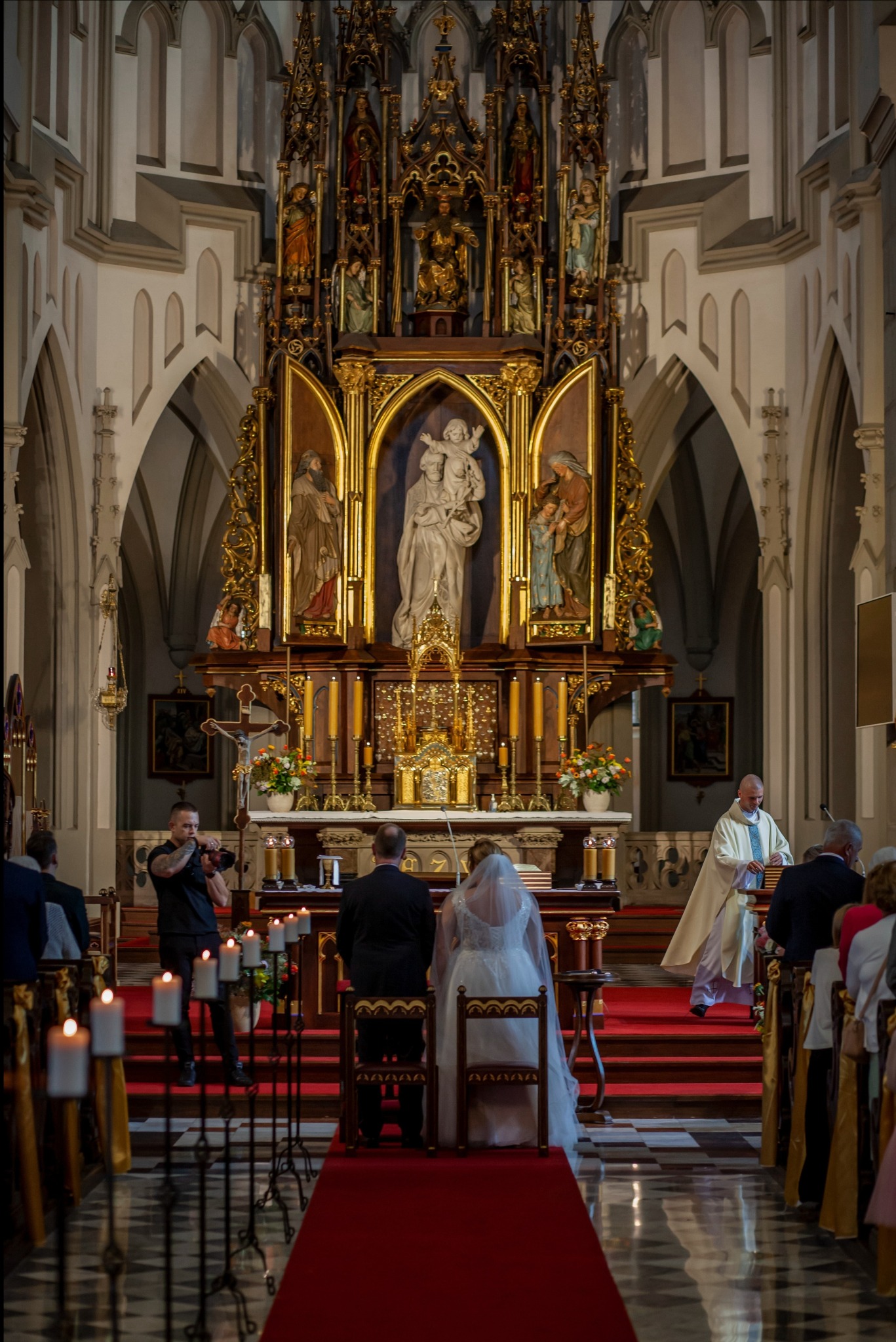 Para młoda podczas ceremonii ślubnej w bogato zdobionym kościele. W tle ołtarz, a po bokach goście. Skrzypek gra na instrumencie. Widok zza pleców pary.