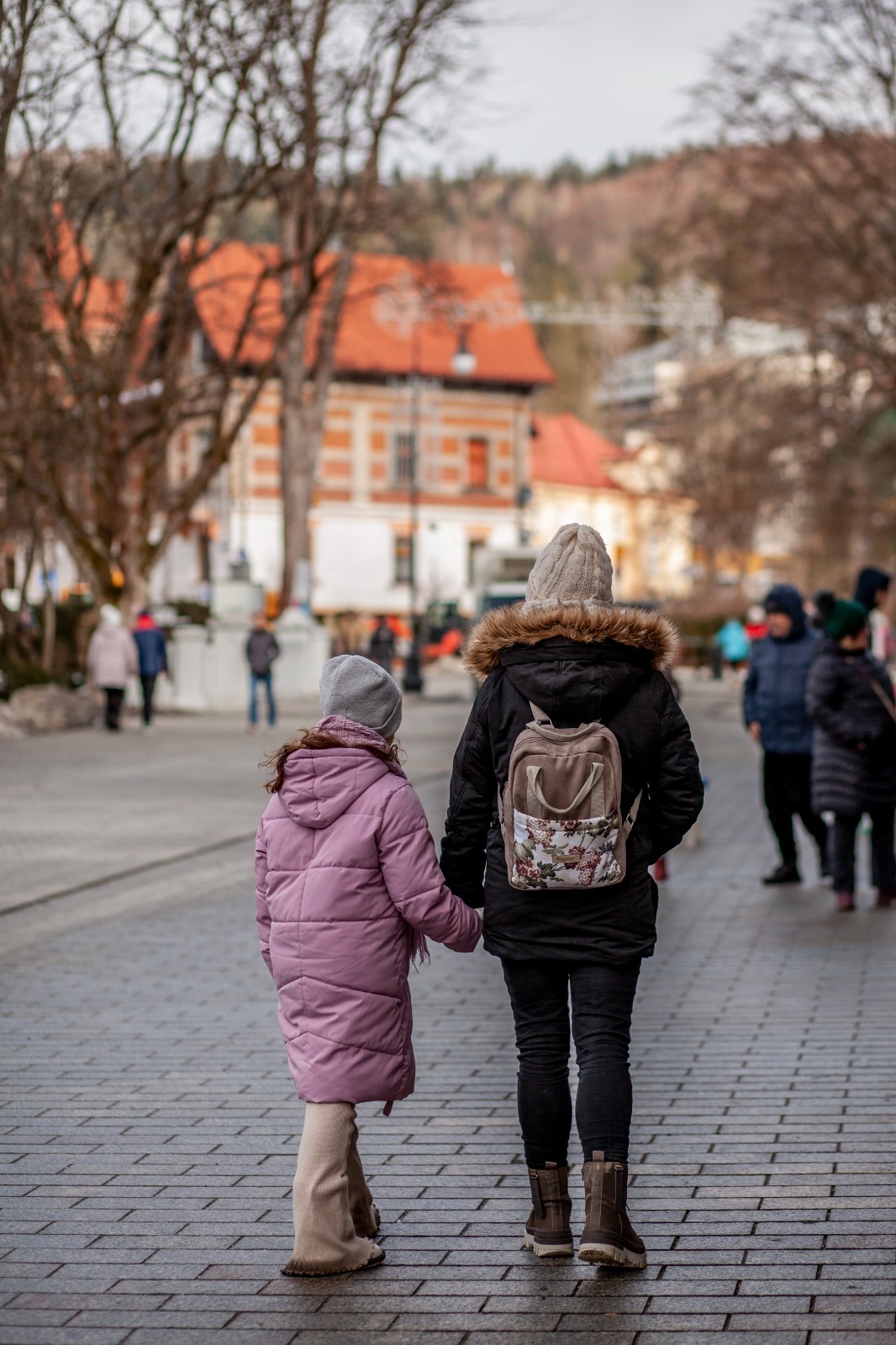Matka z córką spacerują po brukowanej ulicy, trzymając się za ręce. W tle budynki z czerwonymi dachami i drzewa bez liści. Ujęcie z perspektywy od tyłu.