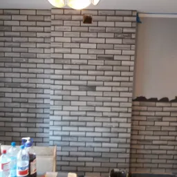 Ściana wewnętrzna wykończona szarą cegłą dekoracyjną, widoczne niedokończone prace wykończeniowe i przygotowanie do malowania ściany obok.