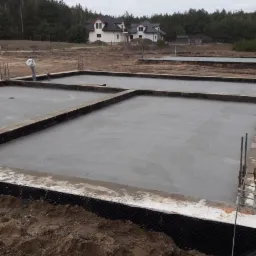 Wylane fundamenty pod budowę domu jednorodzinnego, widoczne zbrojenia i izolacja przeciwwilgociowa, w tle zarys zabudowy mieszkalnej i las.