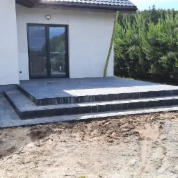 Nowoczesny dom z wyjściem na taras wyłożonym szarą kostką brukową, widoczne schody prowadzące do drzwi tarasowych, w tle iglaki i fragment dachu sąsiedniego budynku.