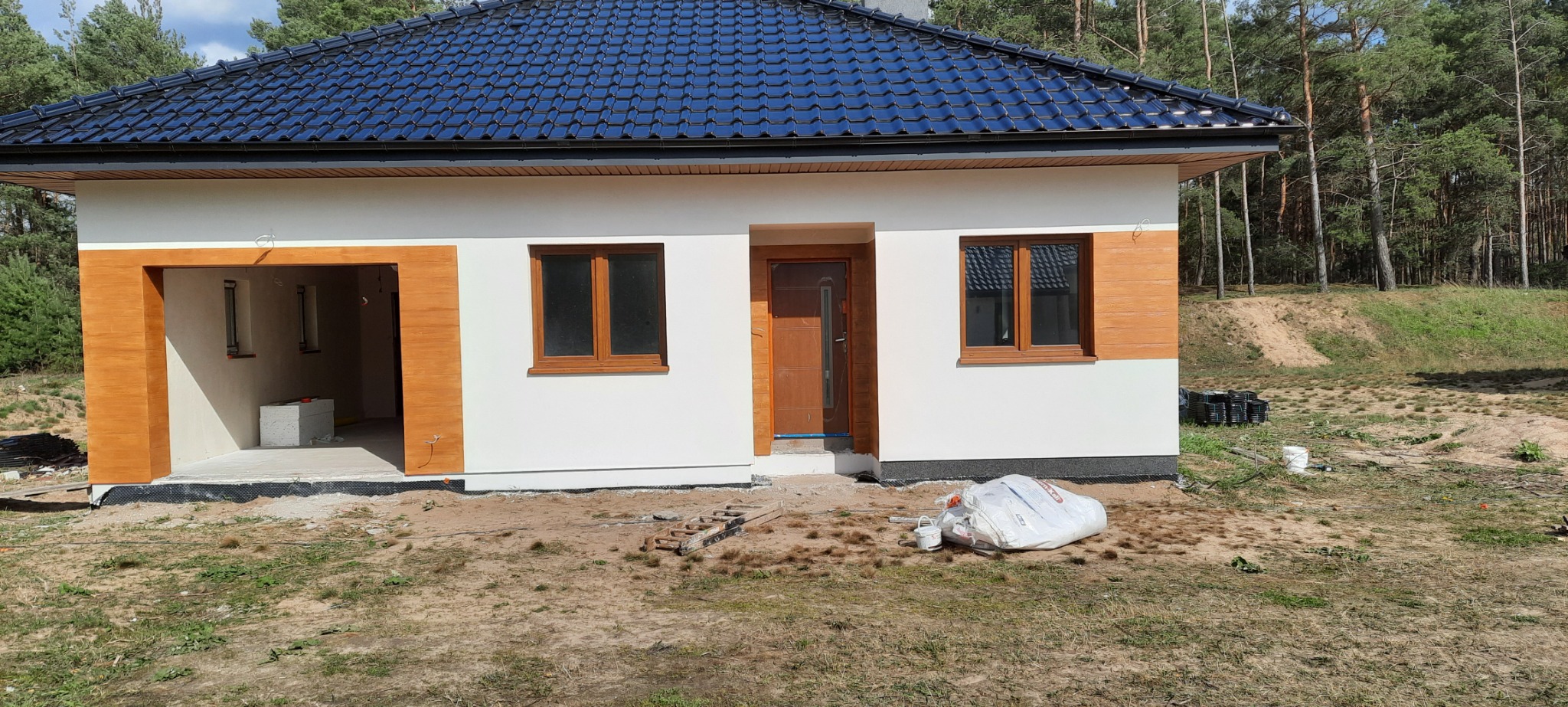 Nowo wybudowany dom jednorodzinny z białymi ścianami i drewnopodobnymi detalami elewacji, widoczne okna z brązowymi ramami i ciemna dachówka, teren wokół domu w trakcie prac wykończeniowych.