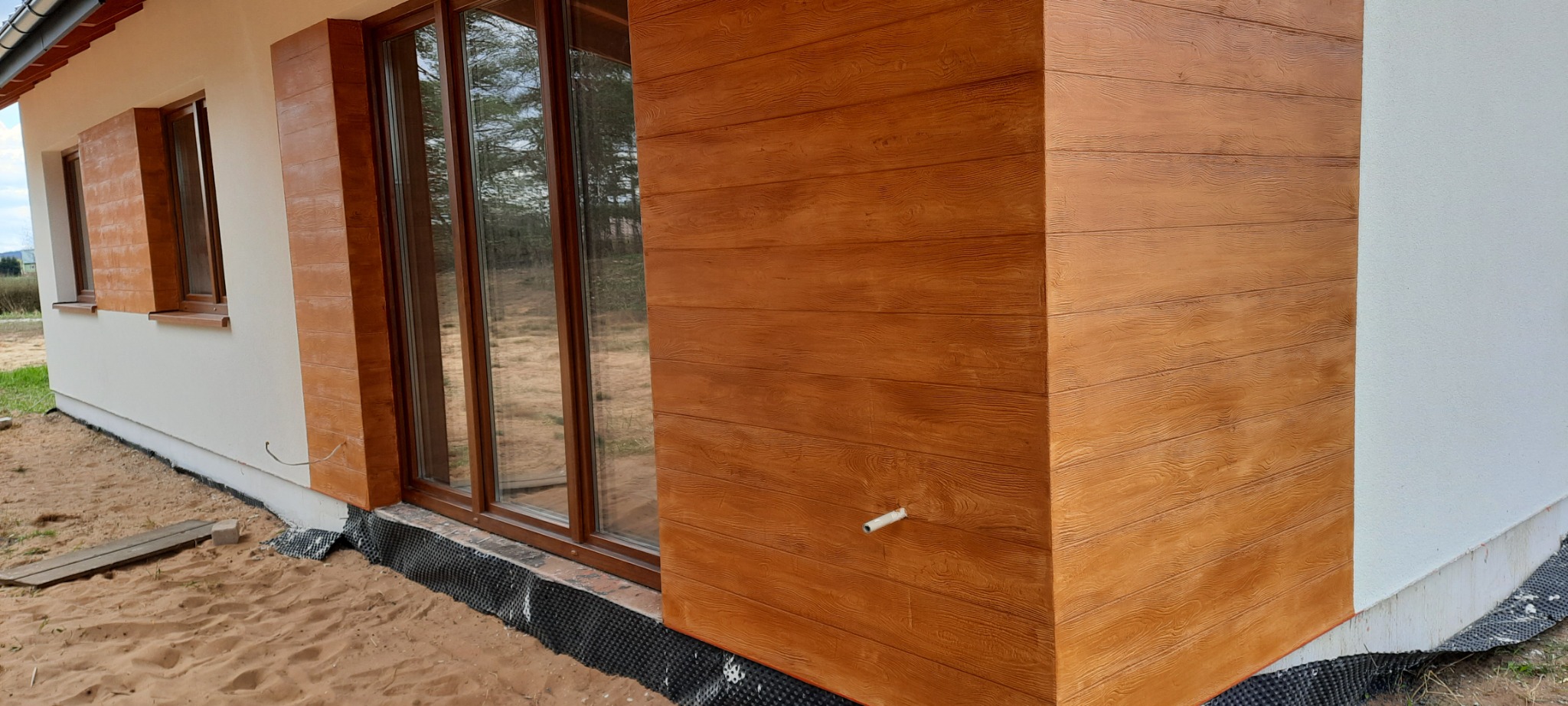 Nowoczesny dom z elewacją imitującą drewno, brązowe okna i białe ściany, widoczne detale wykończenia.