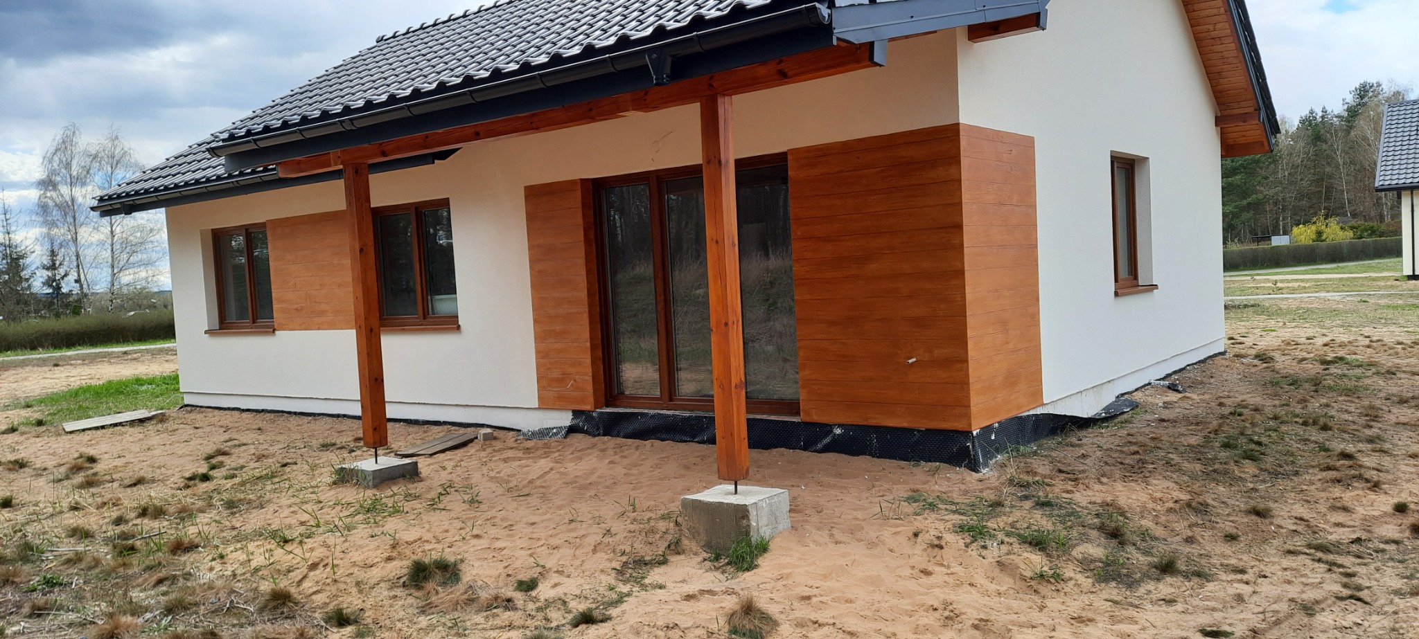Nowoczesny dom z elementami drewnianej elewacji, widoczne fundamenty zabezpieczone folią, drewniane słupy podtrzymujące zadaszenie nad tarasem, surowy teren wokół budynku.