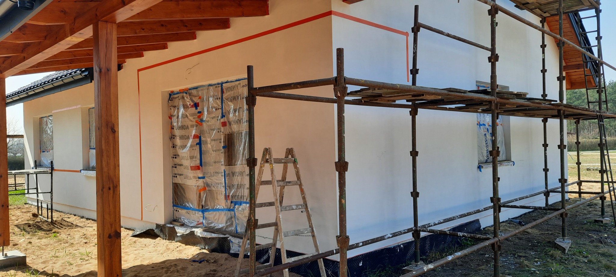 Dom w trakcie prac elewacyjnych: ściany otynkowane na biało, okna zabezpieczone folią, widoczne rusztowania i drabina, zaznaczone pomarańczową taśmą linie