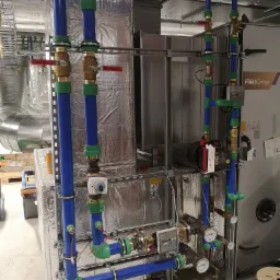 Kompleksowa instalacja rur w kolorze niebieskim i zielonym z zaworami z czerwonymi uchwytami, manometry i czujniki na tle srebrnej izolacji i urządzenia FläktGroup.