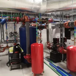 Kompleksowa kotłownia z widocznymi rurami w różnych kolorach, zbiornikami ciśnieniowymi i pompami obiegowymi, z elementami odzieży roboczej i odkurzaczem przemysłowym, ukazująca zaawansowany etap...