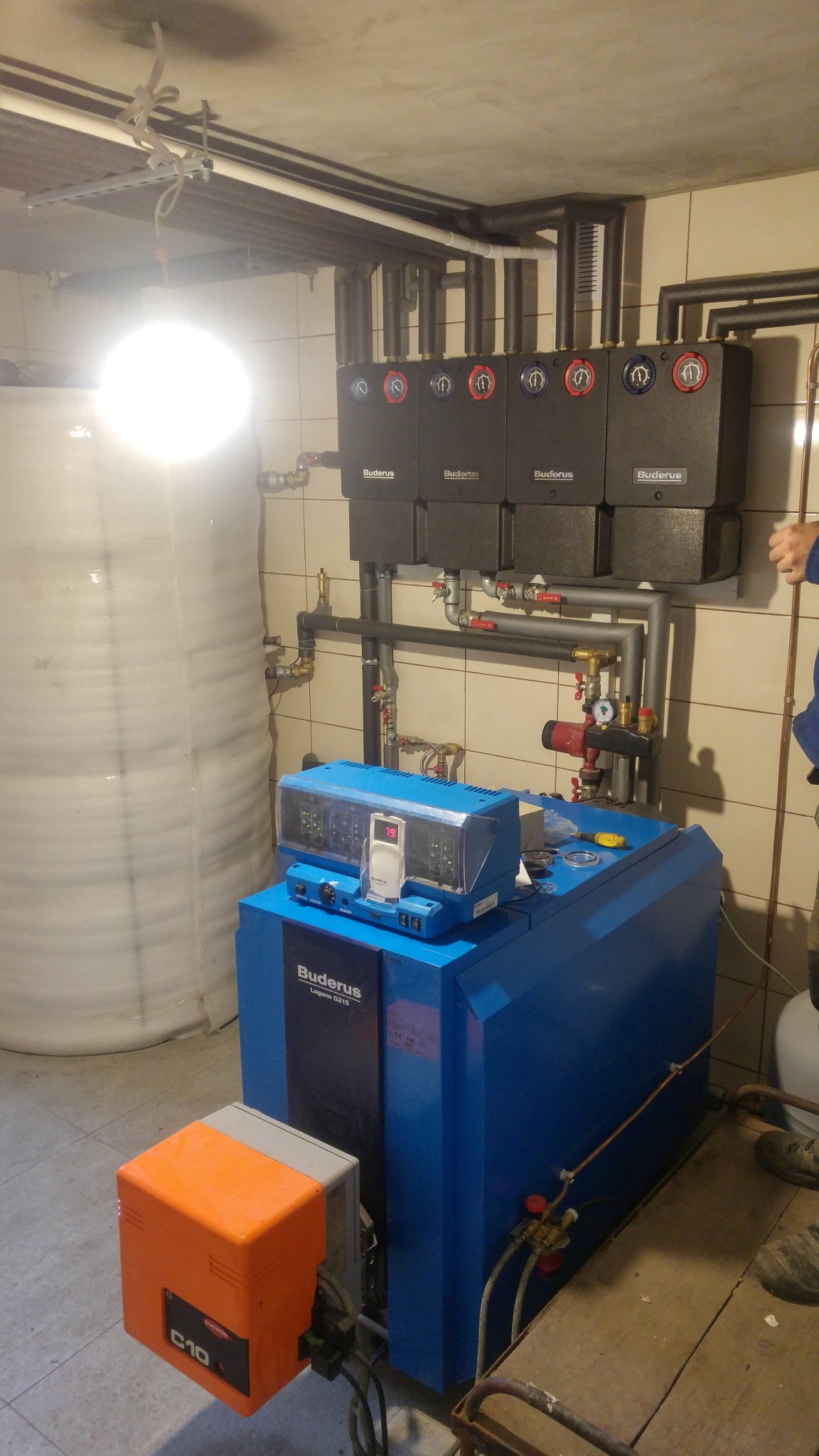 Kompletna kotłownia Buderus z zasobnikiem ciepłej wody, rozdzielaczem i automatyką, z widocznymi rurami instalacji grzewczej i wodnej na tle płytek ceramicznych.