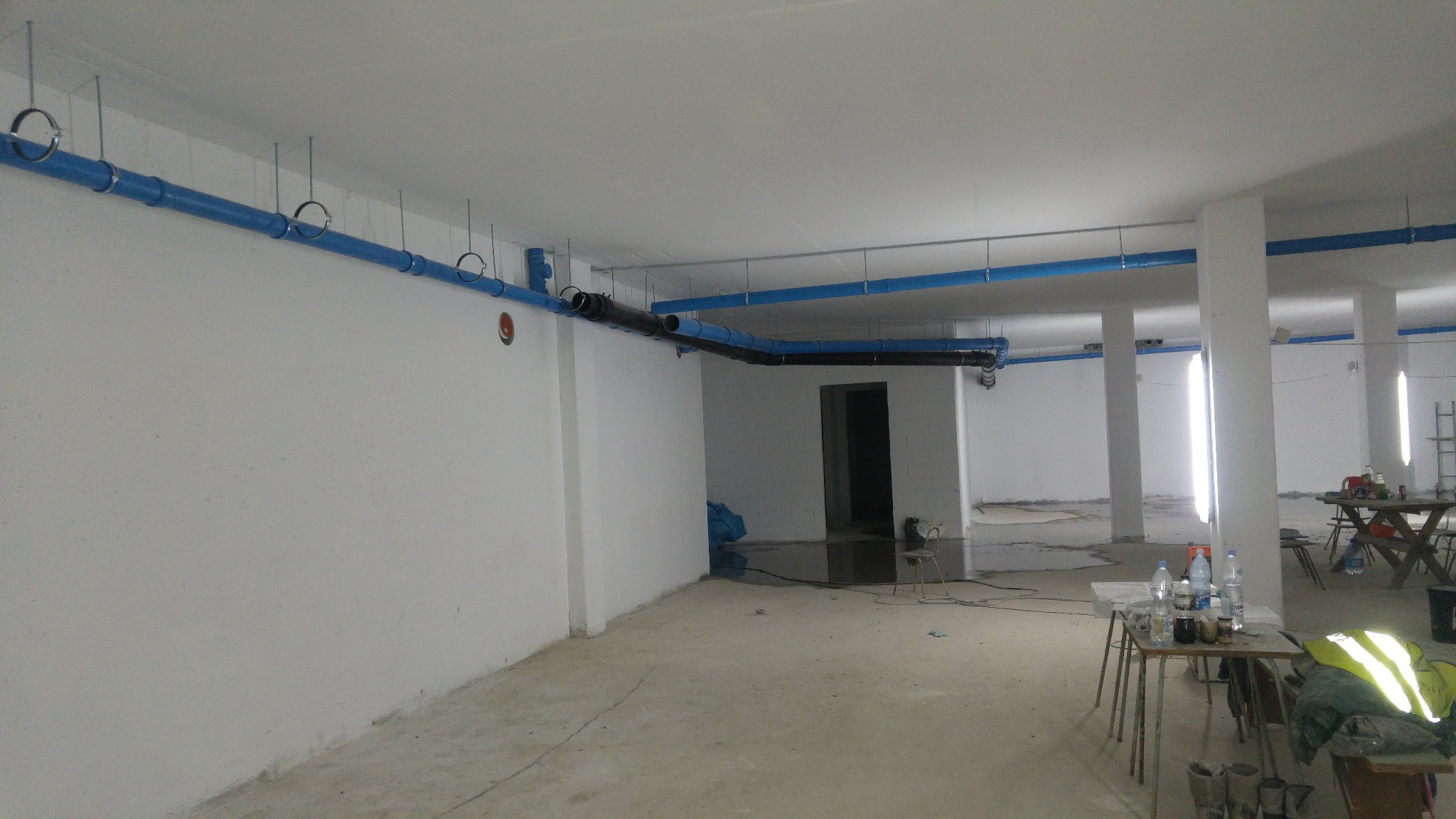 Wnętrze w trakcie prac instalacyjnych: widoczne niebieskie i czarne rury podwieszone pod sufitem, surowy beton na podłodze, białe ściany, stoliki z butelkami wody, odblaskowa kamizelka i robocze buty.