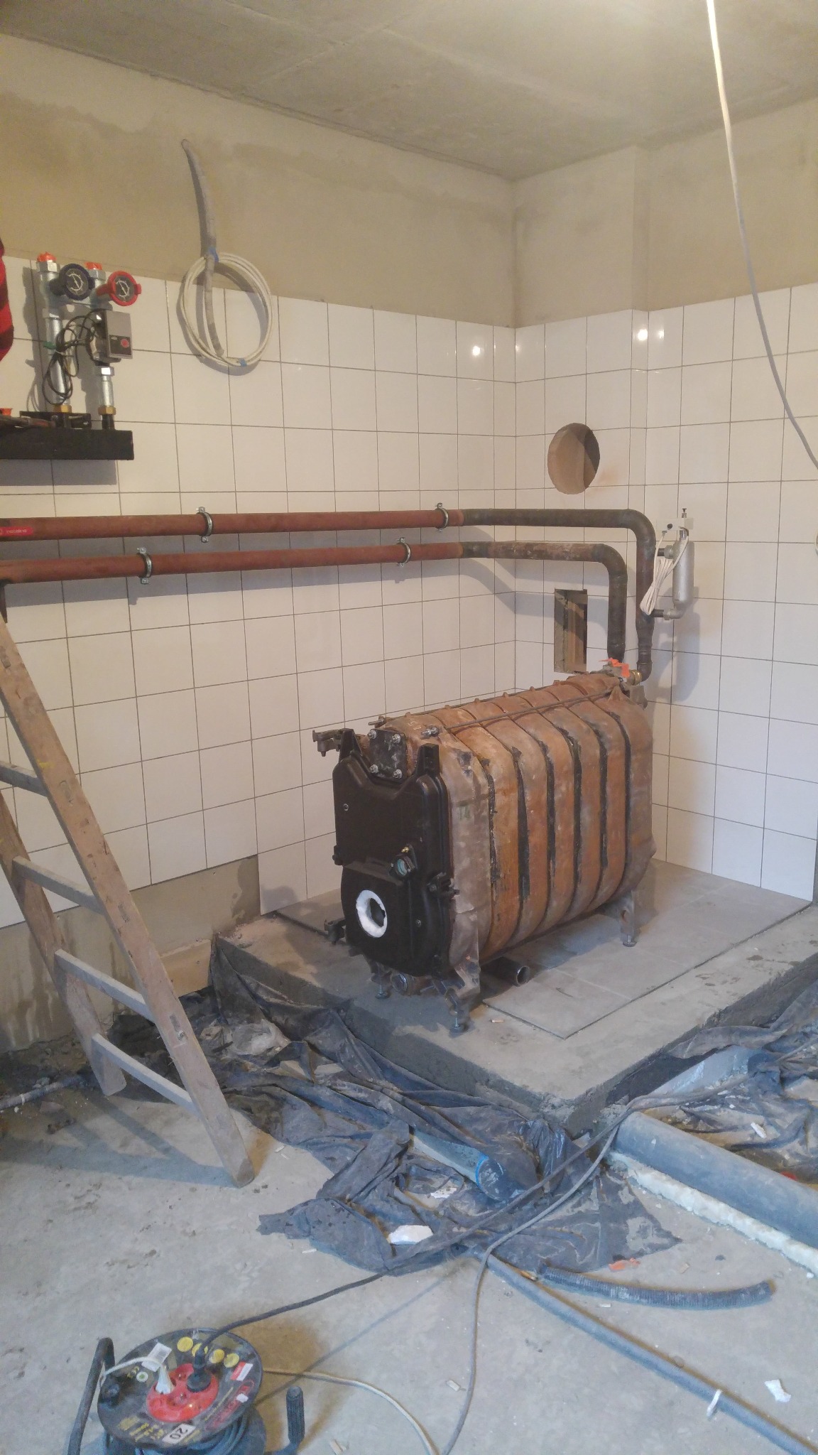 Montaż pieca centralnego ogrzewania w pomieszczeniu z wyłożonymi kafelkami ścianami, widoczne rury, drabina i rozłożona folia ochronna na podłodze.