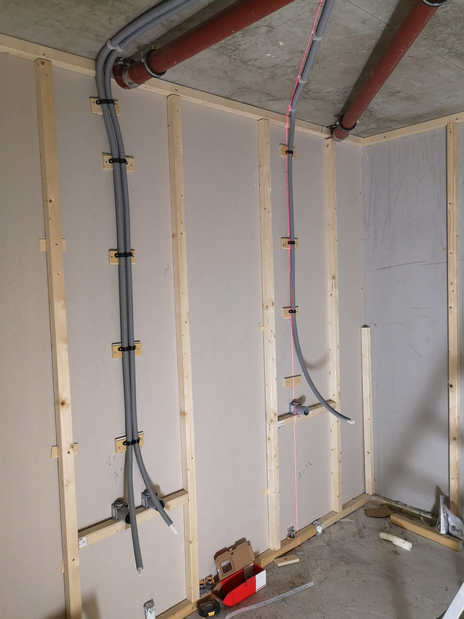 Wewnętrzne ściany w trakcie budowy z widocznymi rurami instalacyjnymi mocowanymi do drewnianej ramy, widoczna poziomica laserowa wyznaczająca pion.