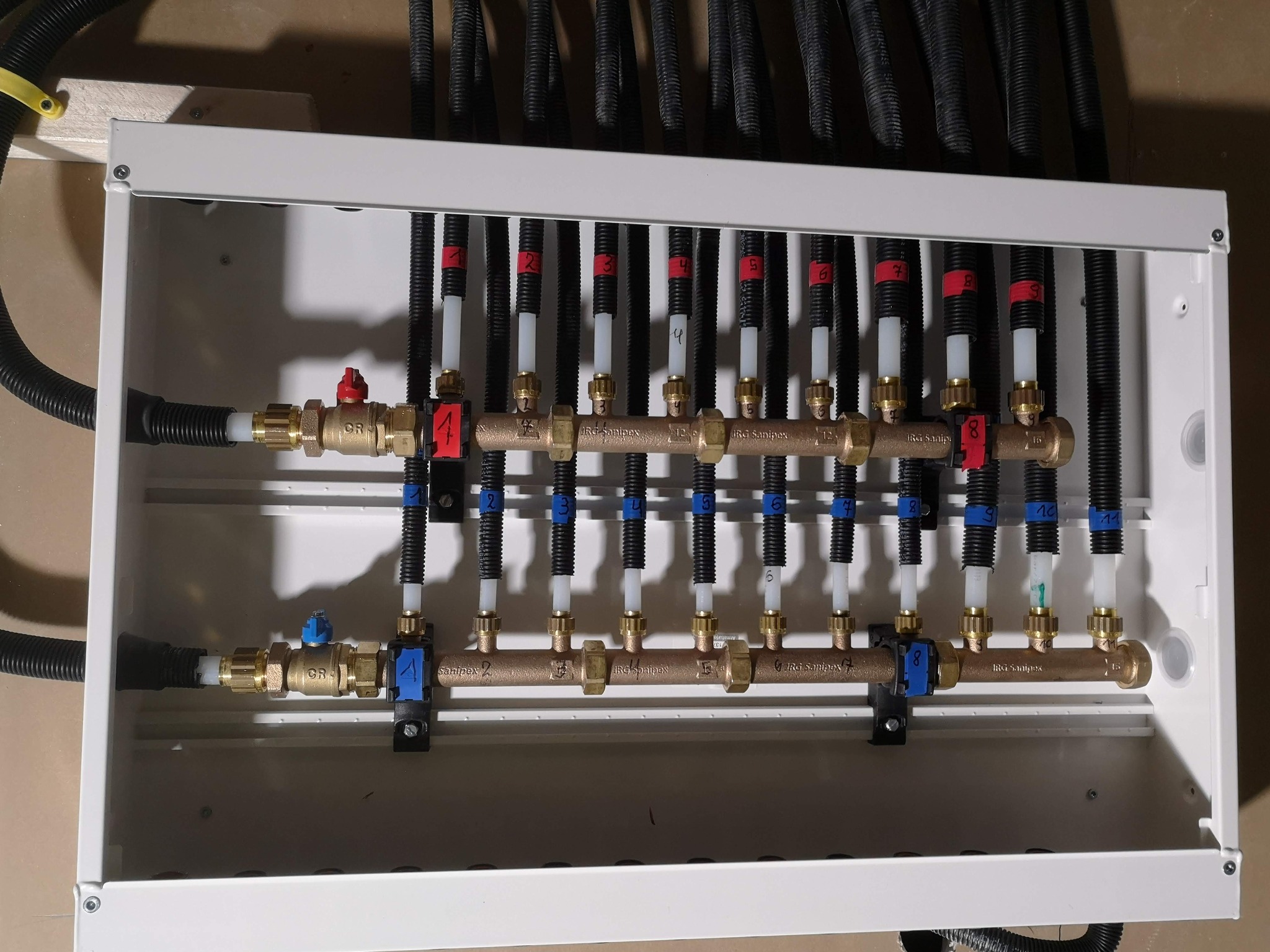 Rozdzielacz instalacji grzewczej w białej skrzynce, z numerowanymi rurami i zaworami, widok z góry pod kątem.
