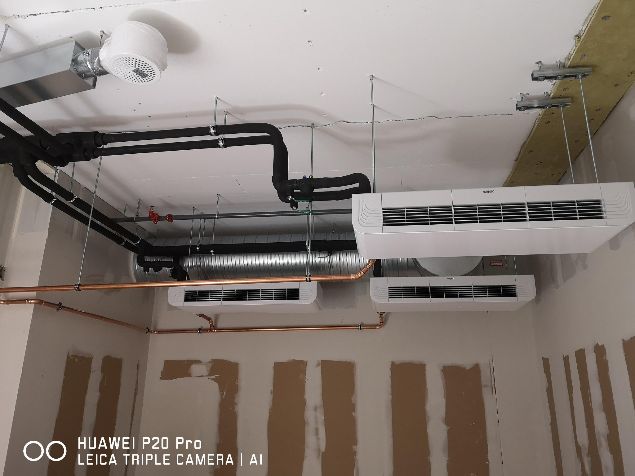 Instalacja grzewcza i klimatyzacyjna w surowym pomieszczeniu: widoczne miedziane rury, czarne izolowane przewody oraz podwieszane jednostki klimatyzacyjne Aermec, sufit z widocznymi elementami...