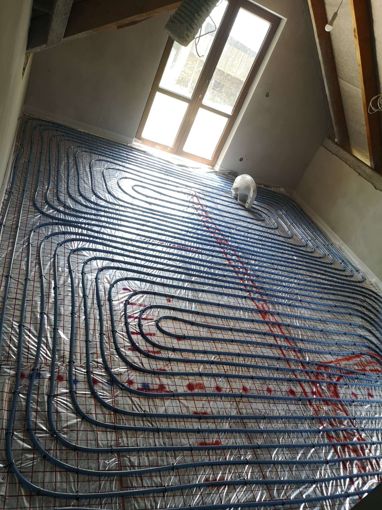 Niedokończona instalacja ogrzewania podłogowego w poddaszowym pomieszczeniu z oknem dachowym; widoczne niebieskie rurki ułożone na folii z czerwoną siatką, przytwierdzone plastikowymi klipsami;...