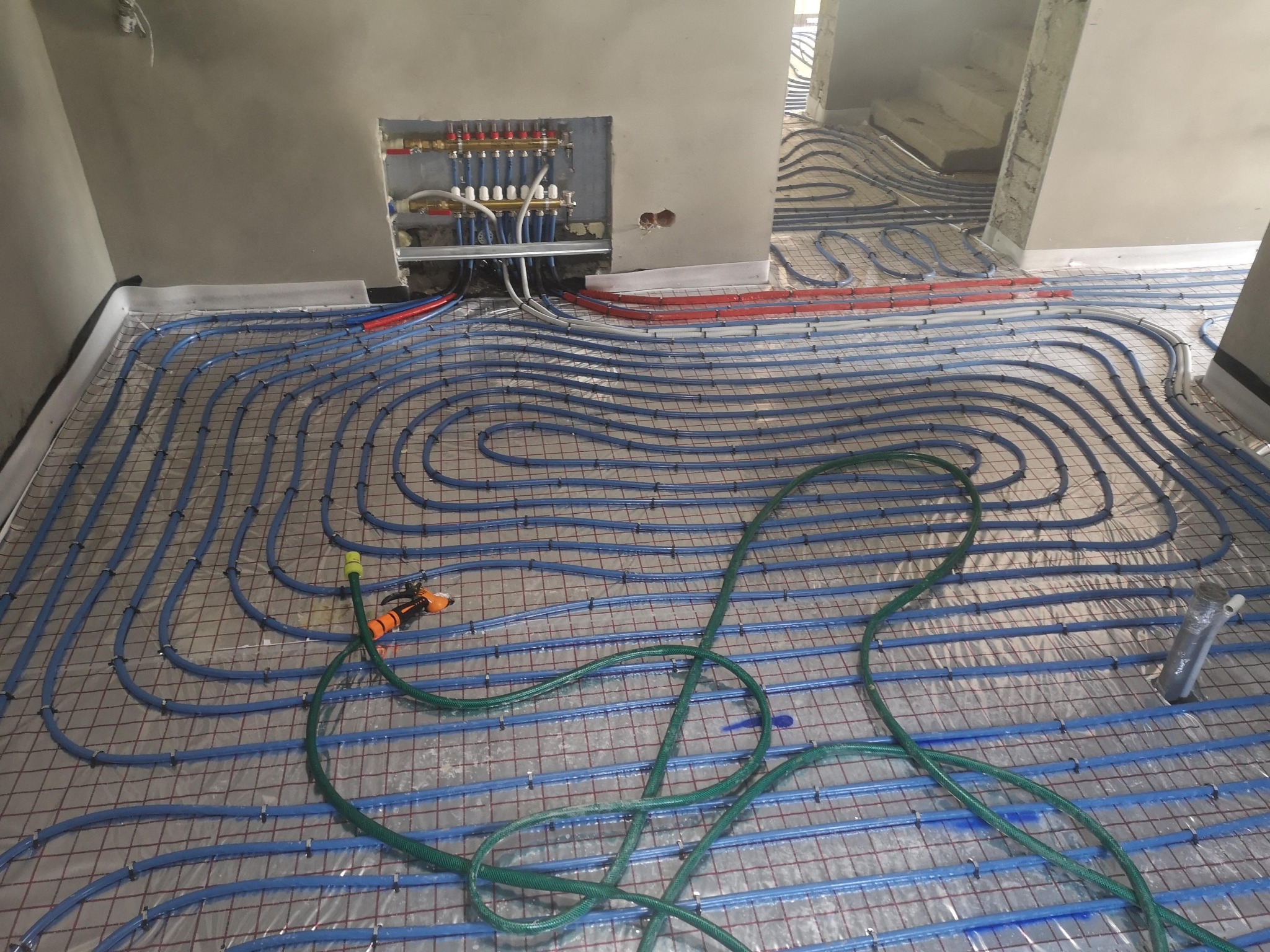 Rozłożona instalacja ogrzewania podłogowego z niebieskich rurek na siatce, widoczny rozdzielacz w ścianie, zielony wąż ogrodowy i elementy instalacji w tle w surowym stanie.