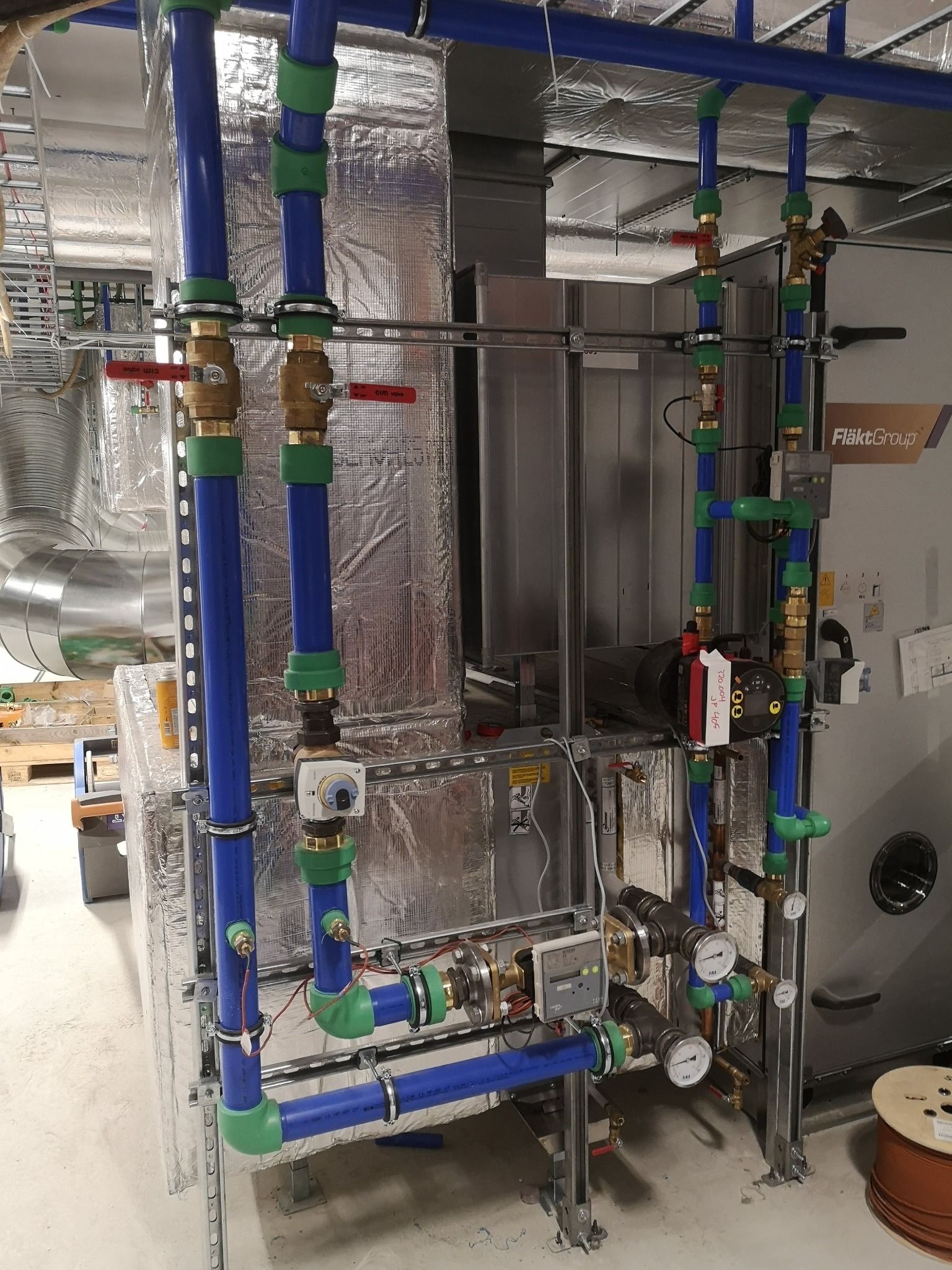 Kompleksowa instalacja rur w kolorze niebieskim i zielonym z zaworami z czerwonymi uchwytami, manometry i czujniki na tle srebrnej izolacji i urządzenia FläktGroup.
