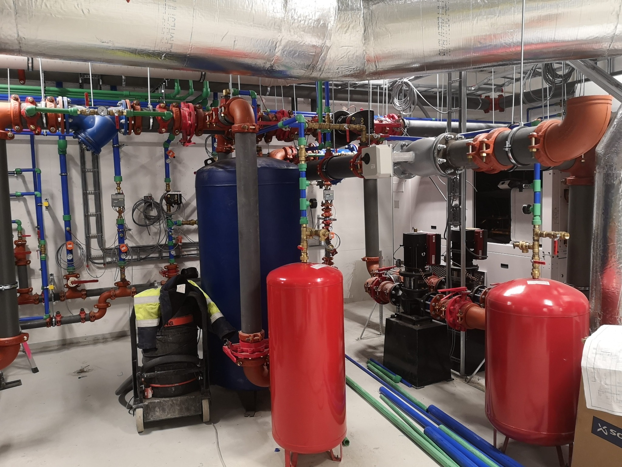 Kompleksowa kotłownia z widocznymi rurami w różnych kolorach, zbiornikami ciśnieniowymi i pompami obiegowymi, z elementami odzieży roboczej i odkurzaczem przemysłowym, ukazująca zaawansowany etap...