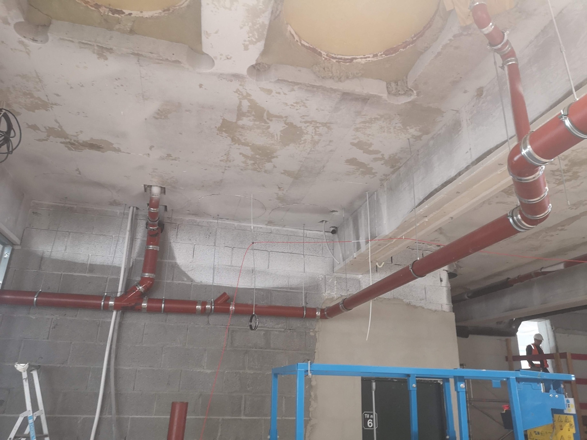 Czerwone rury kanalizacyjne z metalowymi obejmami zamontowane pod surowym, betonowym sufitem w trakcie budowy, widoczny podnośnik i pracownik w oddali.