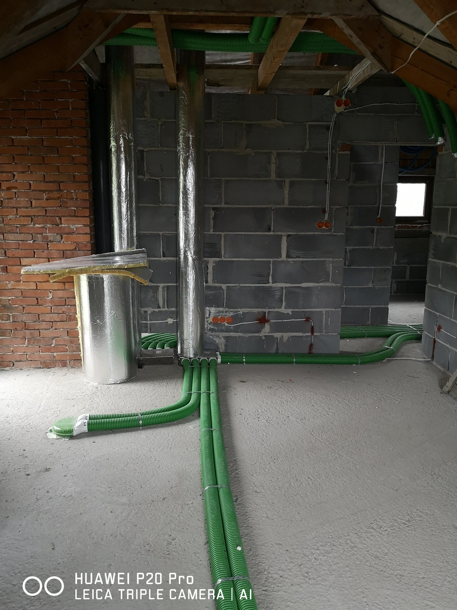Surowy stan poddasza z widocznymi, nowymi instalacjami: pionowe rury w srebrnej izolacji, poziome zielone rury karbowane ułożone na podłodze i podwieszone pod sufitem, fragment ściany z cegły...