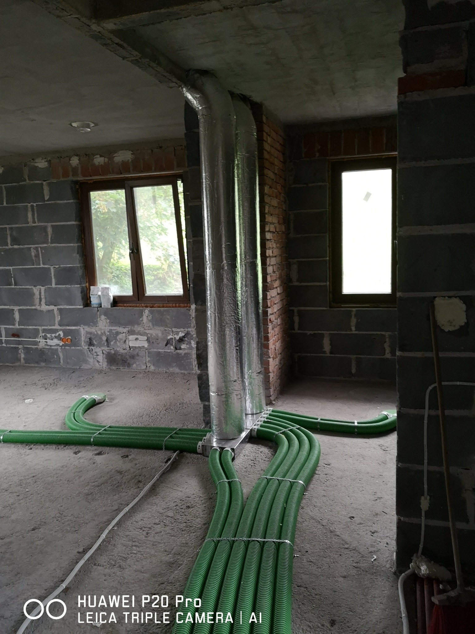 Wnętrze w trakcie budowy z widocznymi zielonymi rurami wentylacyjnymi na podłodze, prowadzącymi do pionowego, srebrnego kanału wentylacyjnego, surowe ściany z cegły i betonu, okna z widokiem...