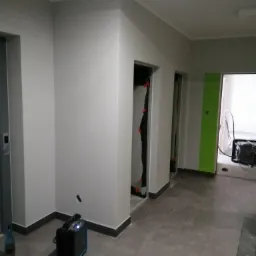 Klatka schodowa w trakcie malowania: świeżo pomalowane szare ściany, widoczne wejścia do mieszkań z zabezpieczeniami na ościeżnicach, na podłodze stoi nagrzewnica i butelka po napoju.