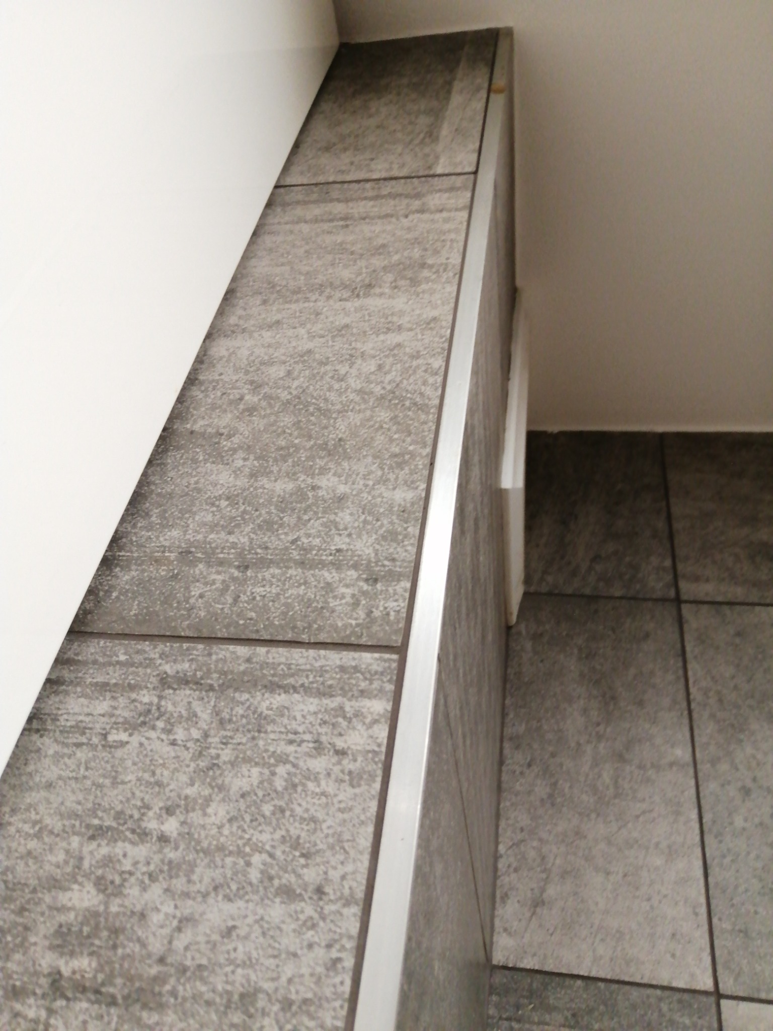 Szare płytki imitujące kamień, ułożone na ścianie i podłodze pod skosem, wykończone metalową listwą, widoczne narożniki i fragment białej ściany.