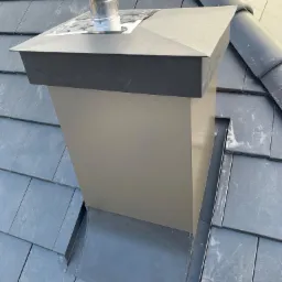 Szary komin wentylacyjny z metalową obudową na dachu pokrytym ciemnymi płytkami łupkowymi, widoczne elementy mocujące i obróbki blacharskie.