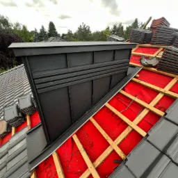 Montaż dachu: Widok z góry na częściowo pokryty dach z widoczną czerwoną membraną, drewnianą konstrukcją łat i stosami szarych dachówek oraz obróbką komina z blachy w kolorze grafitowym.