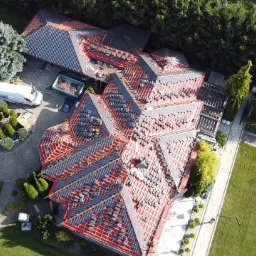 Widok z lotu ptaka na rozległy dach w trakcie układania szarej dachówki na czerwonej membranie, z widocznymi elementami więźby dachowej, na tle zadbanego ogrodu z pojazdami i meblami ogrodowymi.