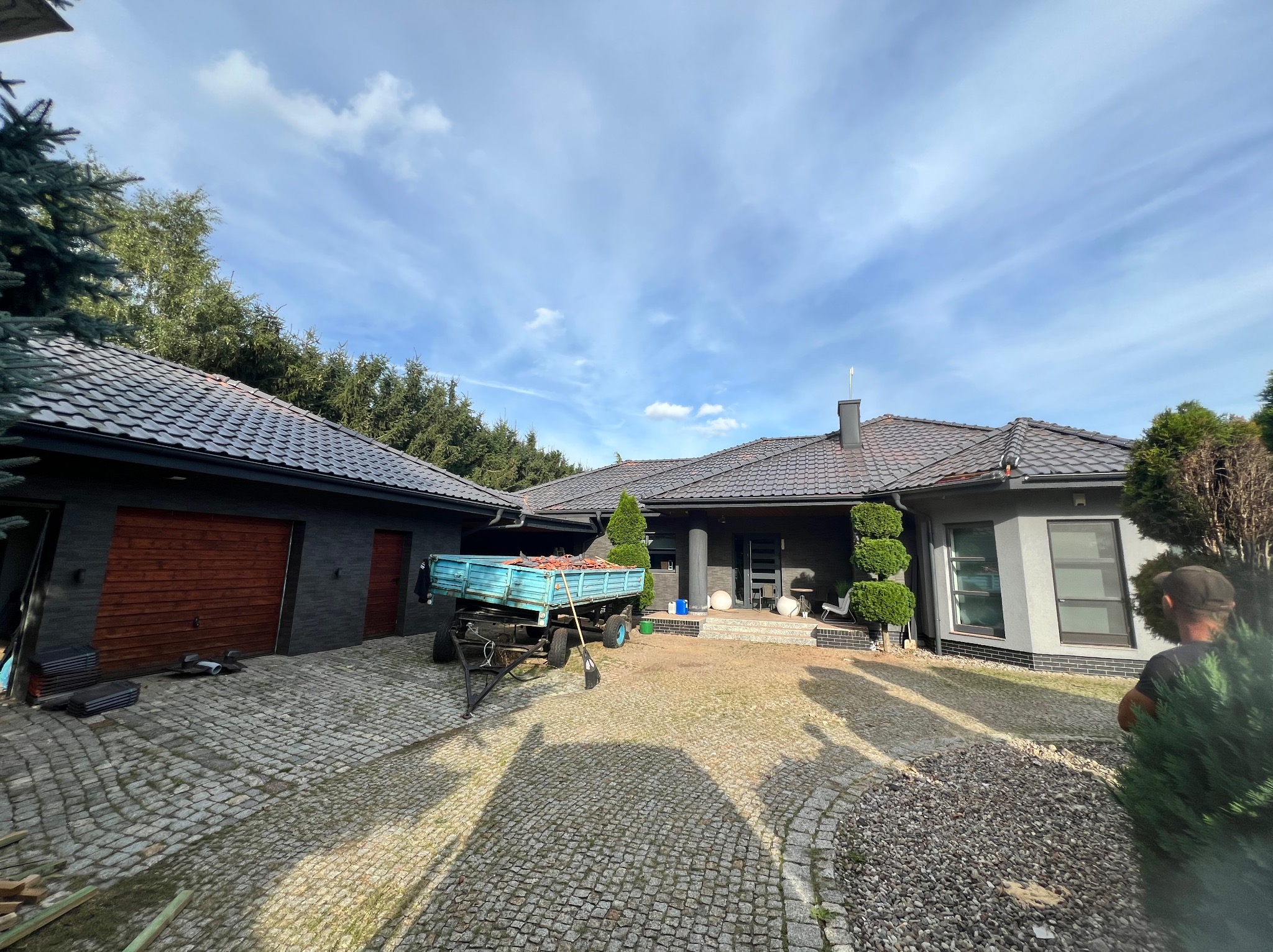 Nowoczesny dom jednorodzinny z ciemną dachówką, widoczny z poziomu brukowanego podjazdu, przyczepa z dachówkami i narzędzia dekarskie sugerują prace w toku.