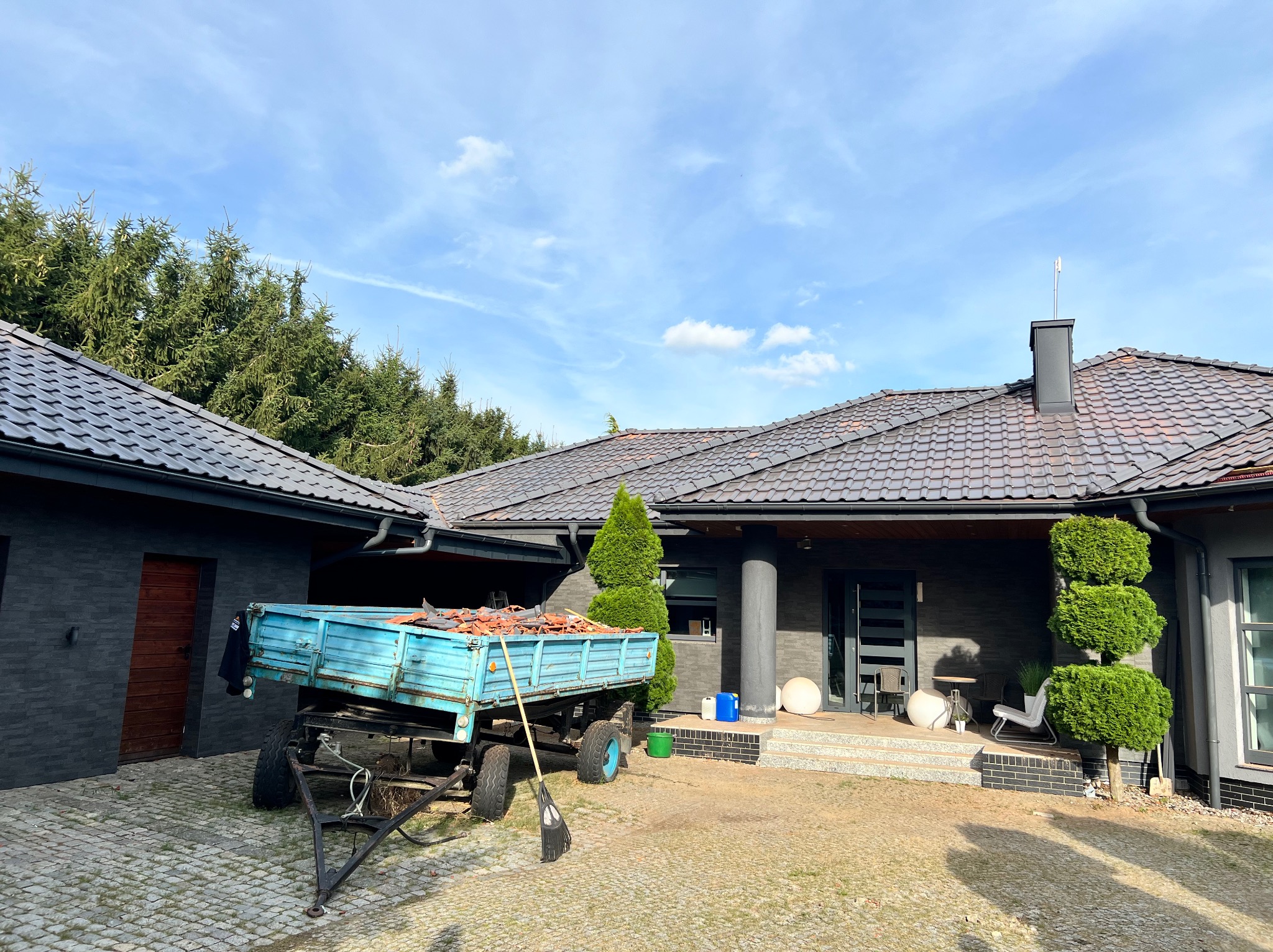 Przyczepa z potłuczonymi dachówkami na tle domu z ciemną dachówką i czarnym kominem, obok leżą narzędzia.