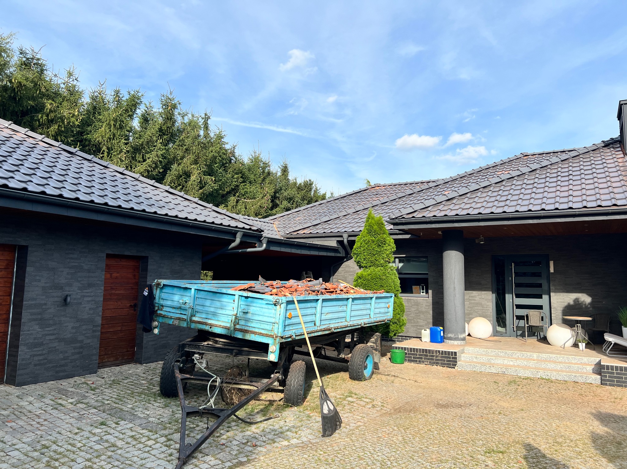 Przyczepa z niebieską skrzynią załadowana starymi dachówkami stoi przed nowoczesnym domem z grafitową elewacją i ciemną dachówką ceramiczną. Na przyczepie oparta miotła. Widoczny fragment dachu...