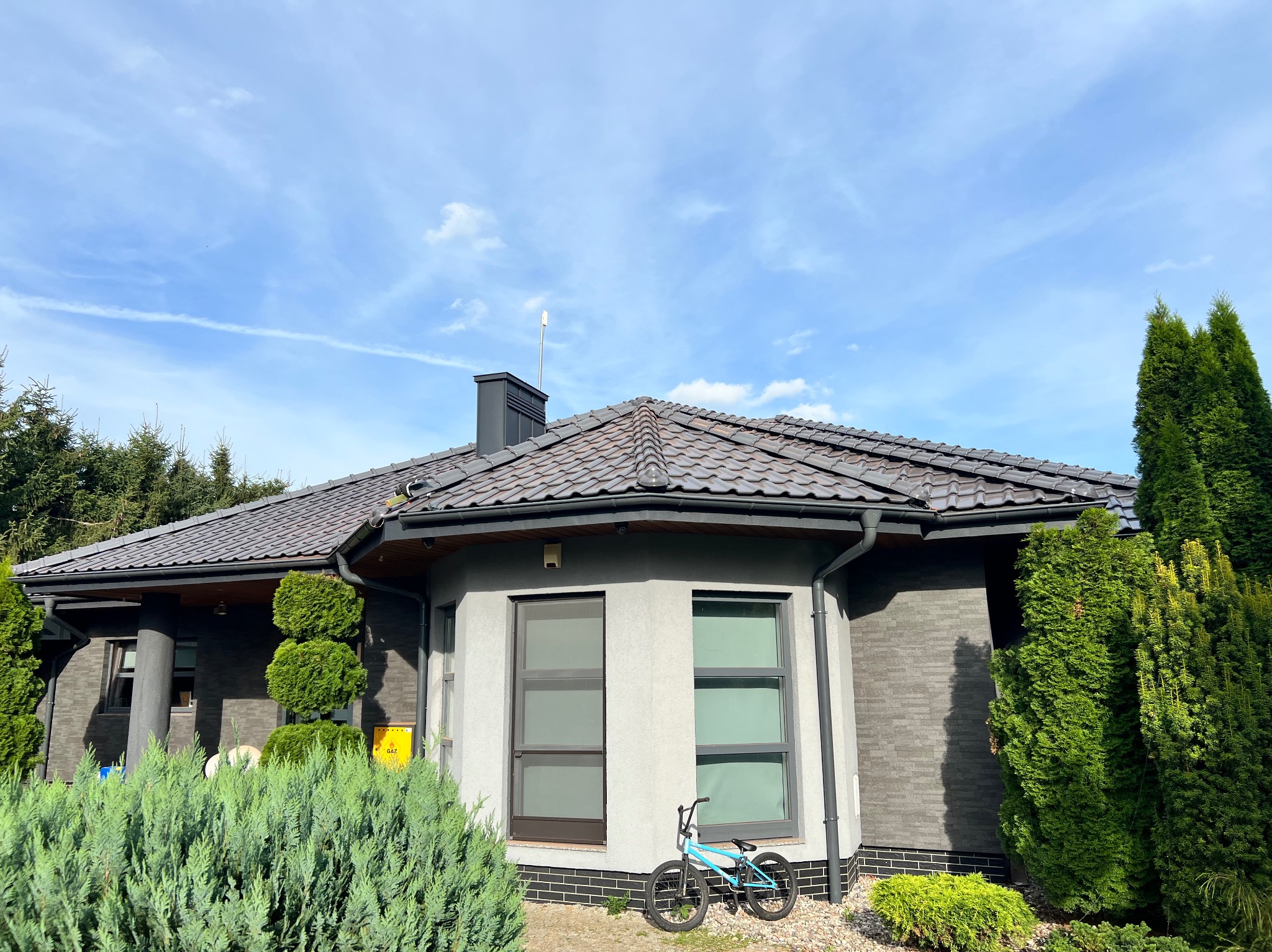 Nowoczesny dom jednorodzinny z ciemną dachówką karpiówką, szarą elewacją z elementami imitującymi cegłę i błękitnym rowerem BMX opartym o ścianę.