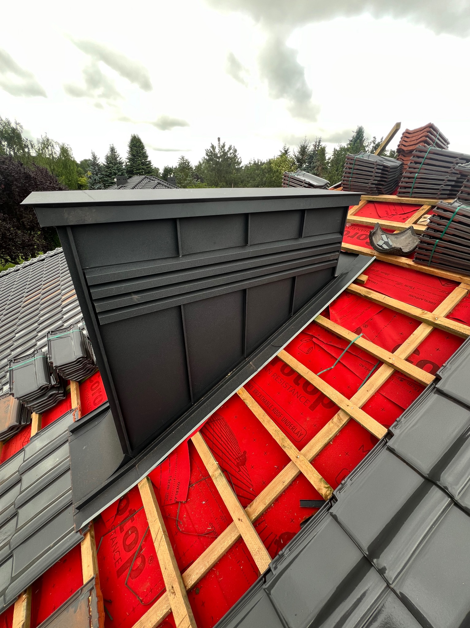 Montaż dachu: Widok z góry na częściowo pokryty dach z widoczną czerwoną membraną, drewnianą konstrukcją łat i stosami szarych dachówek oraz obróbką komina z blachy w kolorze grafitowym.